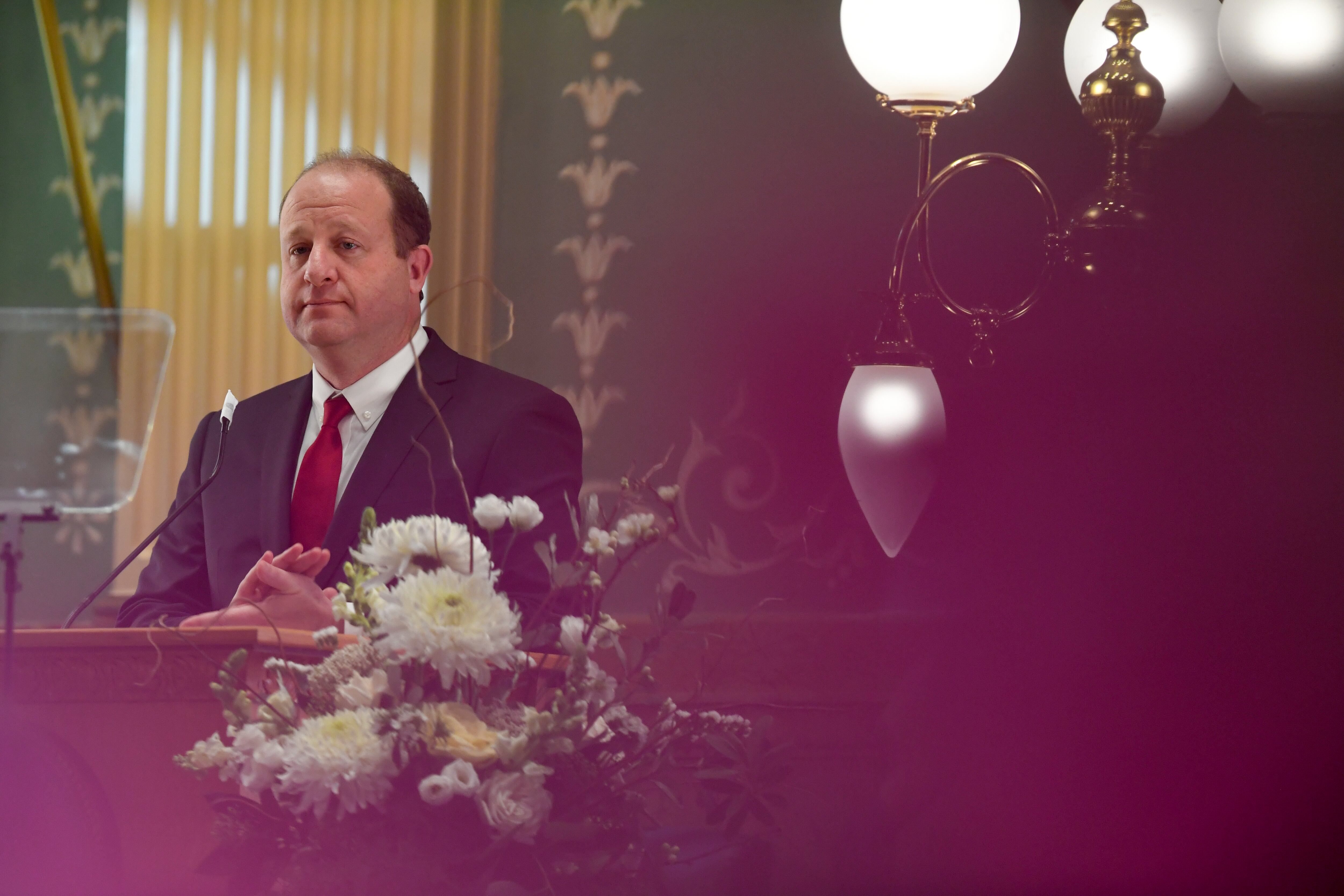 Gov. Jared Polis, in a suit and tie, stands behind a lectern adorned with flowers at the state Capitol.