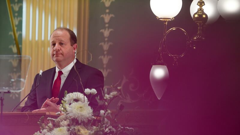 Gov. Jared Polis, in a suit and tie, stands behind a lectern adorned with flowers at the state Capitol.