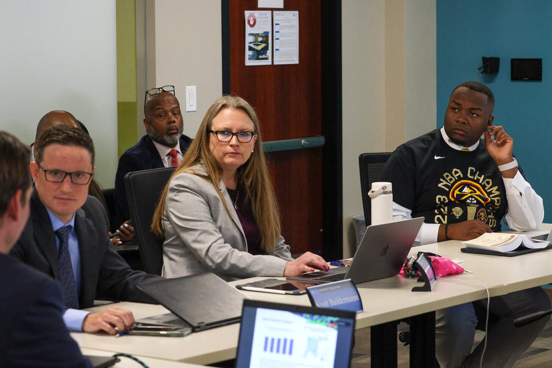 Three people sit at a table with laptops in front of them. There is a person sitting in a chair in the background.