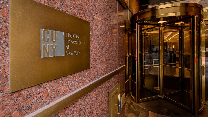 A granite wall with a bronze plaque that reads CUNY The City University of New York. To the right is a revolving door.