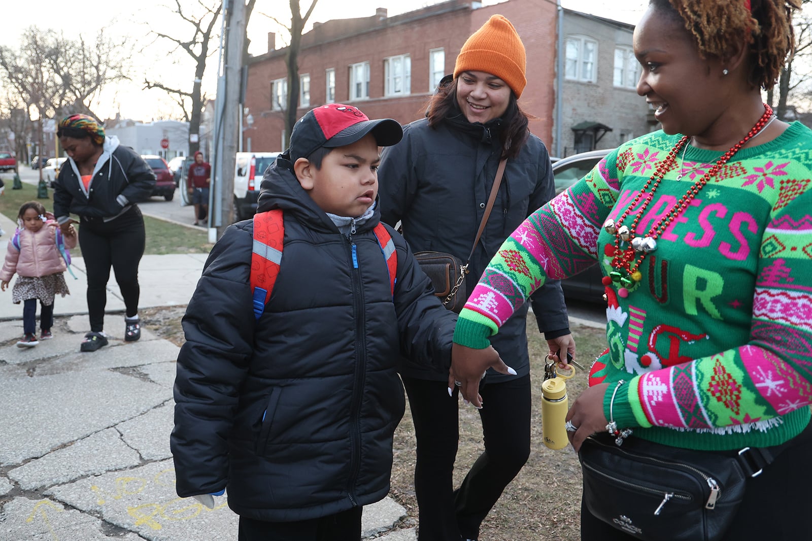 A Black woman in a colorful holiday sweater smiles while grabbing the hand of a child in a black jacket and red hat while his mom in an orange hat stands next to them and smiles.