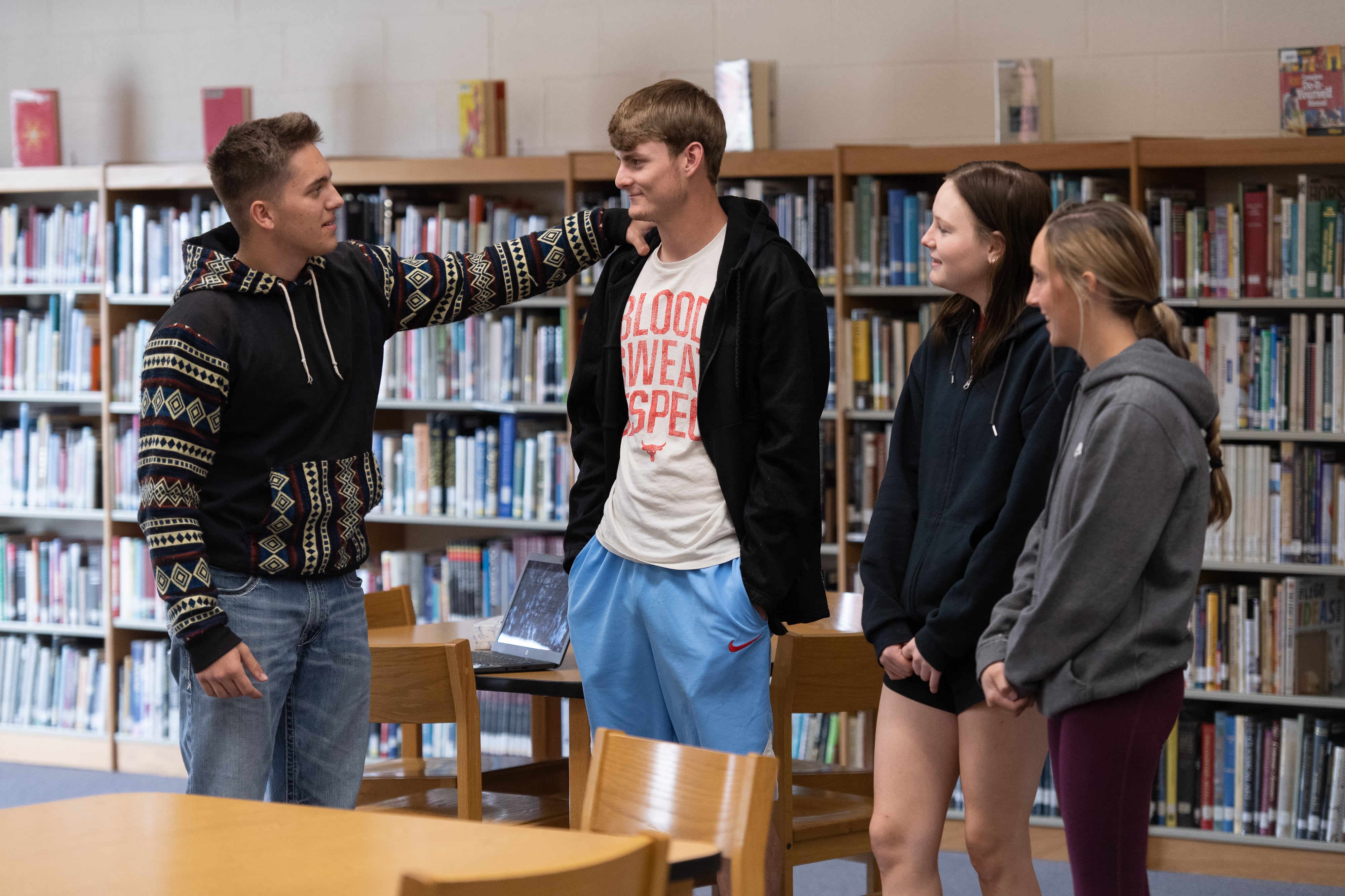 Four high school students stand next to each other talking inside of a school library.