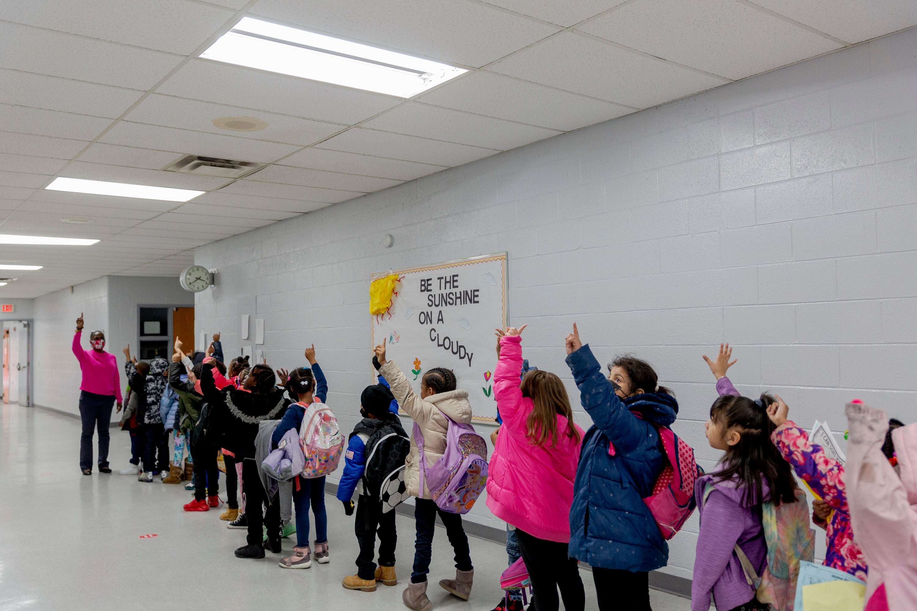 A line of young students wearing their coats and backpacks stand in front of a an adult in a hallway with white brick walls.