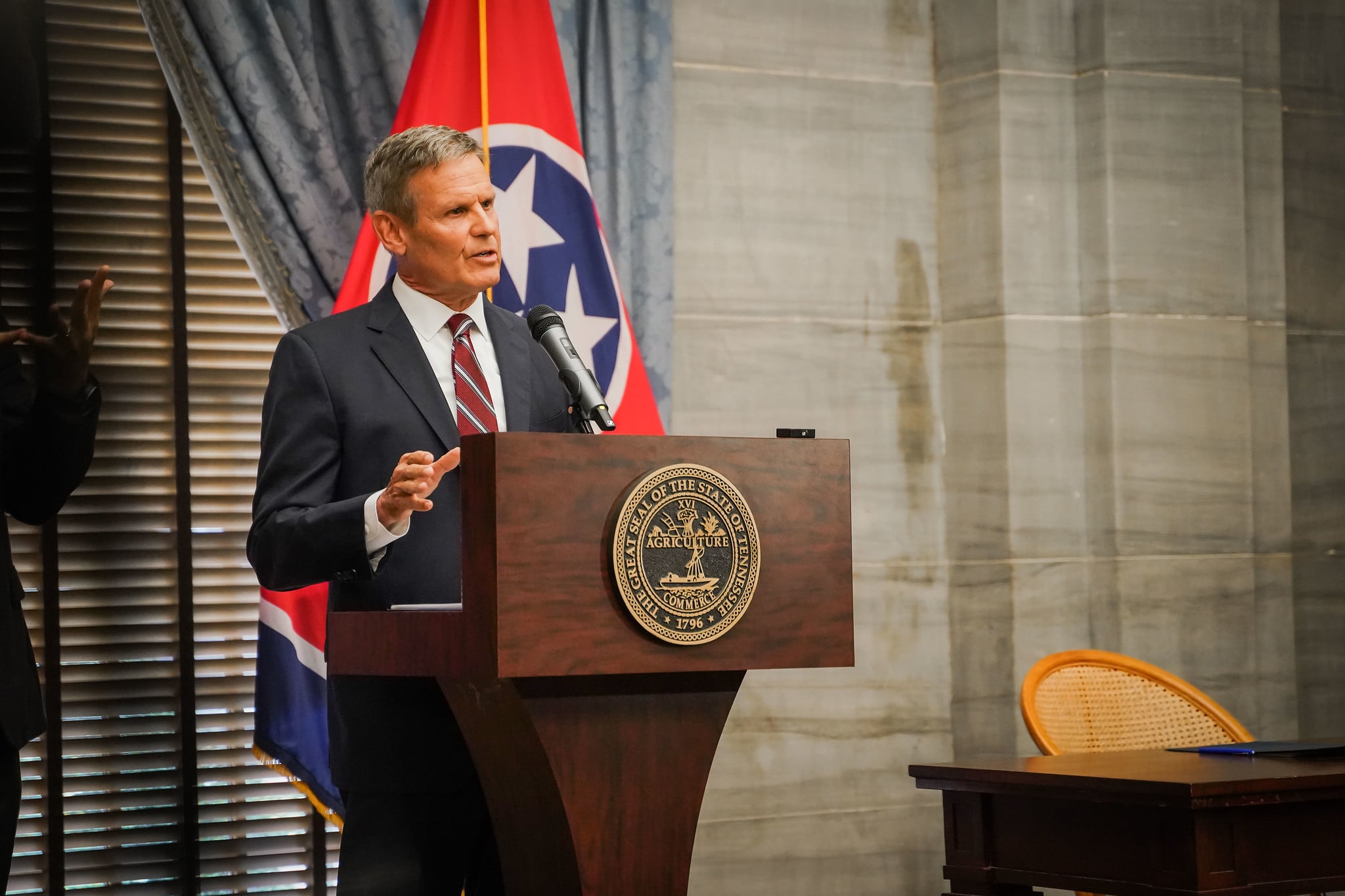 A man wearing a suit and tie speaks at a podium against the backdrop of the Tennessee state flag.