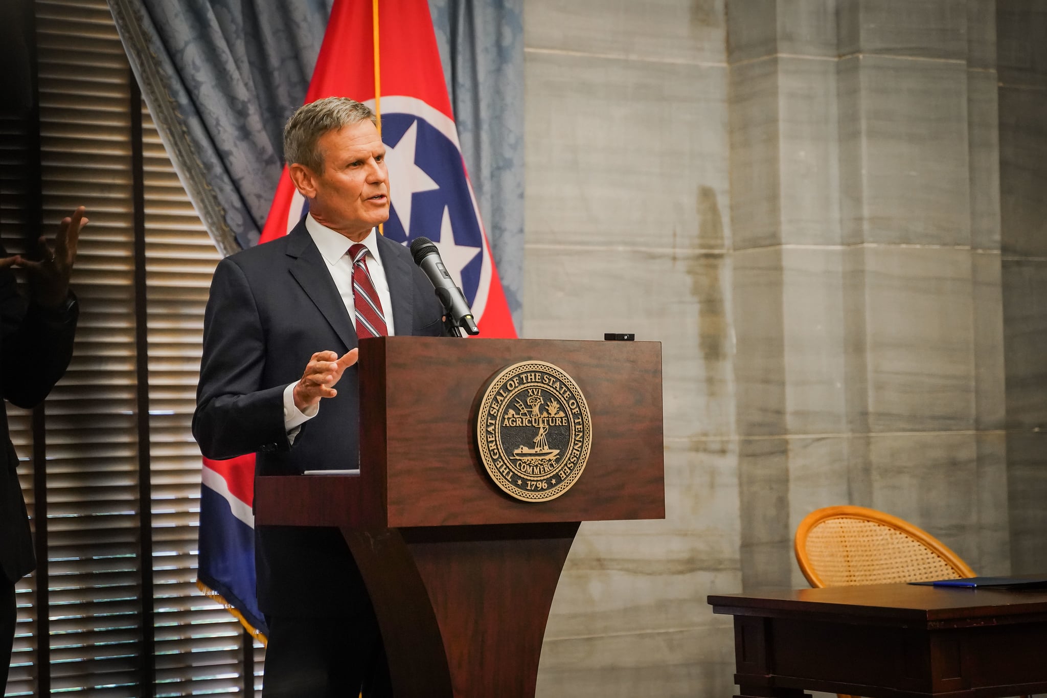 A man wearing a suit and tie speaks at a podium against the backdrop of the Tennessee state flag.