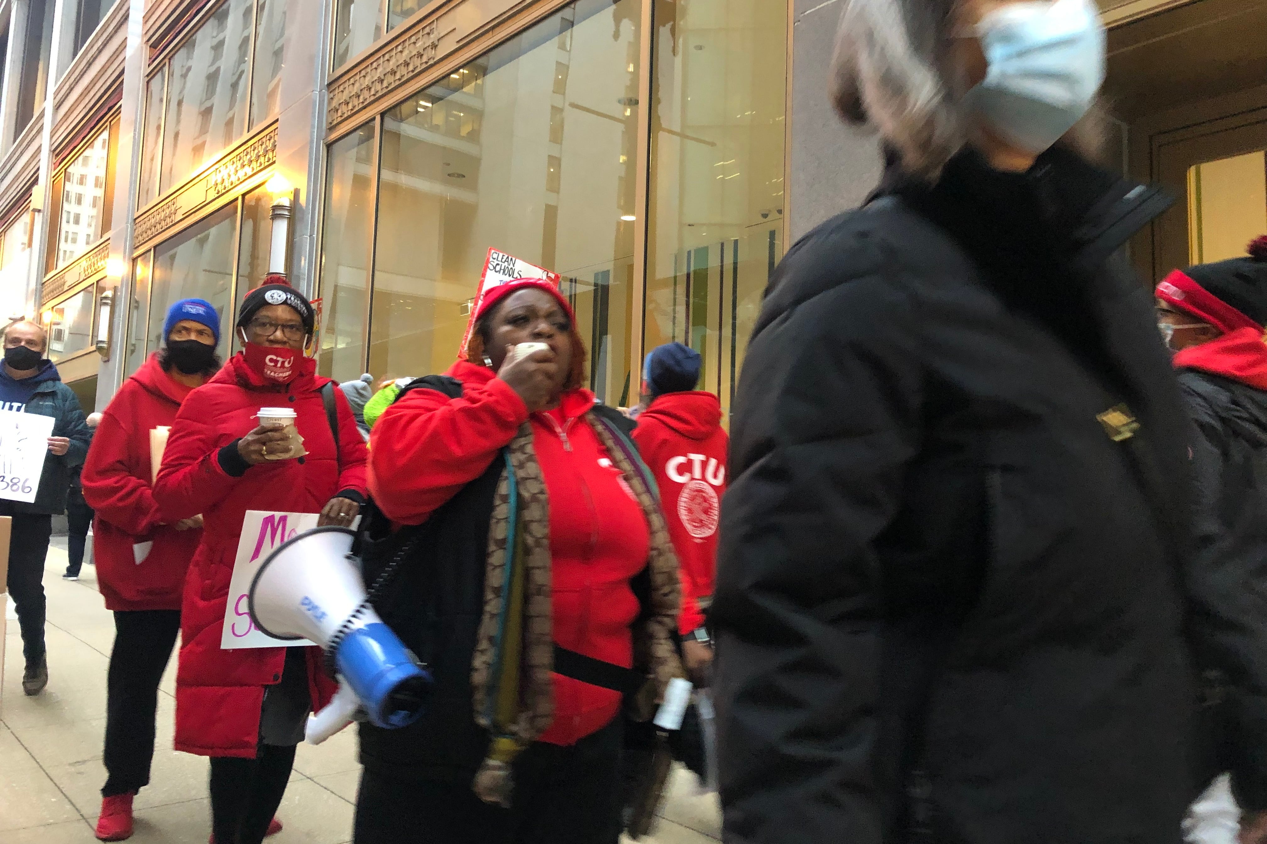 A group of people wearing red carrying posters and a megaphone march on a downtown sidewalk.