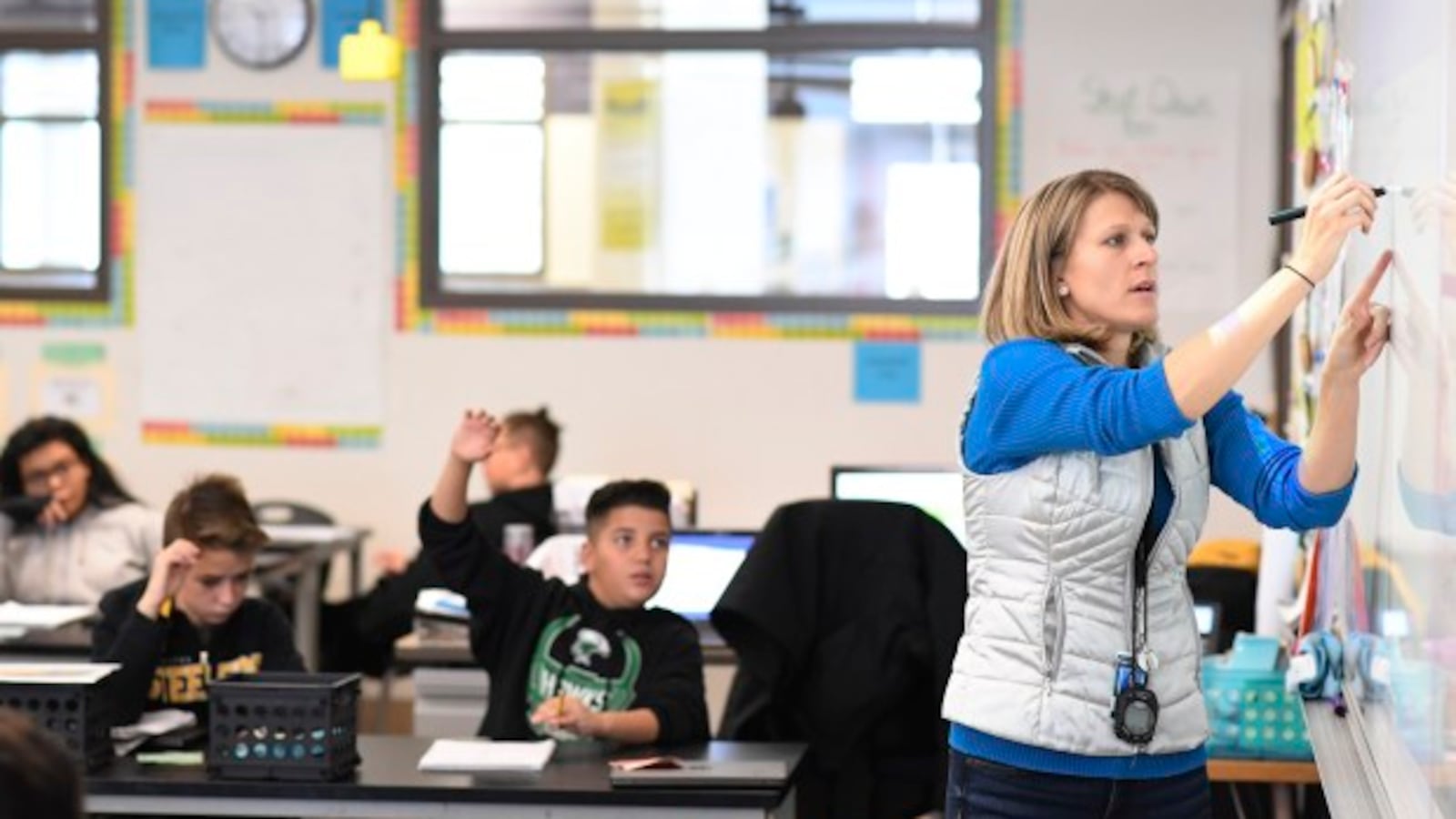A female teacher is writing on a white board to the right as students sit behind her in desks.