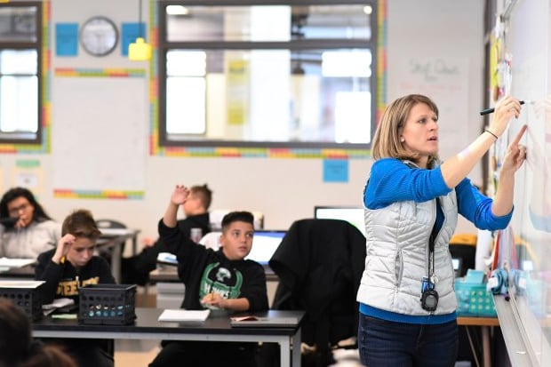 A female teacher is writing on a white board to the right as students sit behind her in desks.