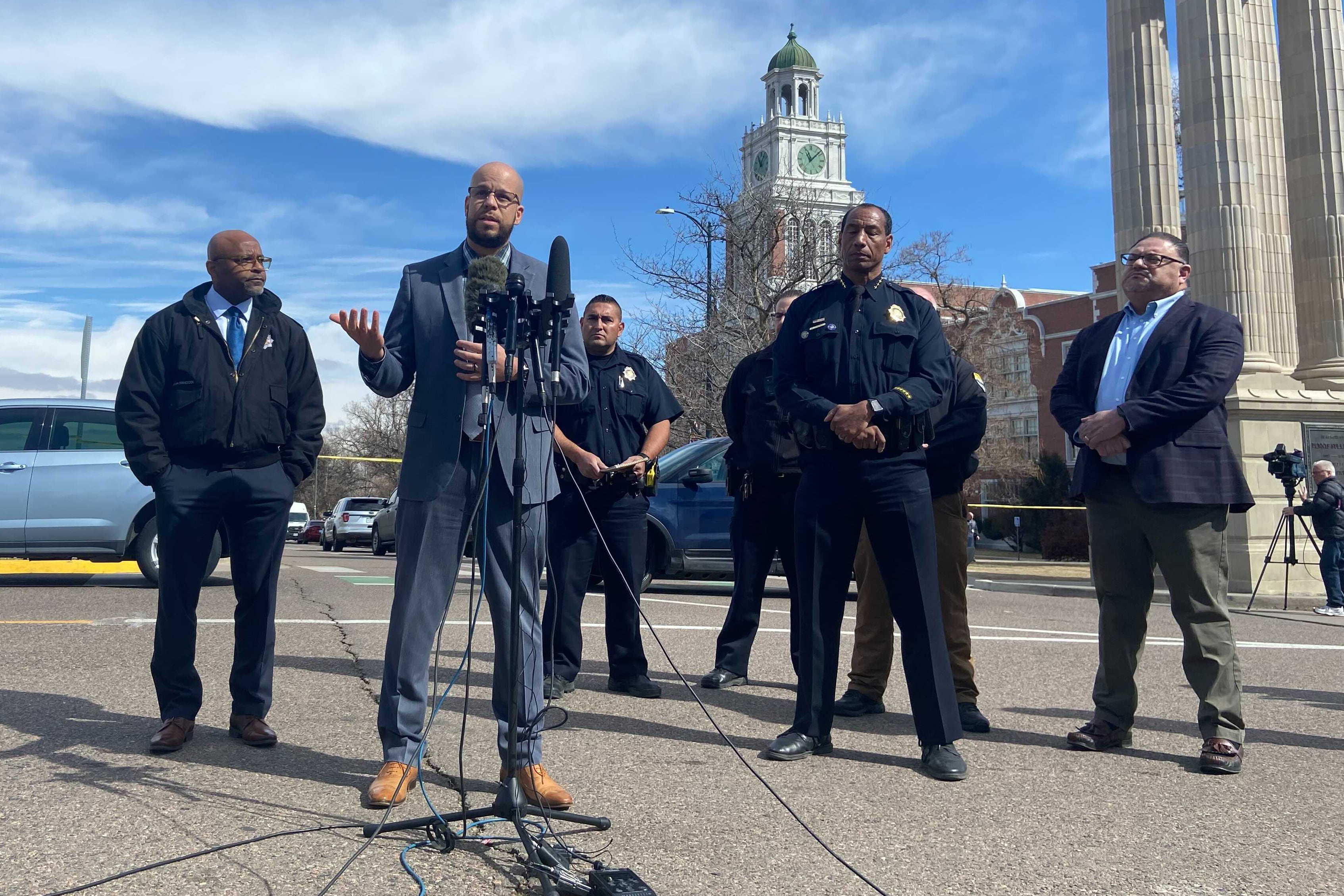 A group of men in suits and one in police dress uniform speak at a press conference. They are outside with a podium and microphones set up in front of them. East High School is visible behind them.