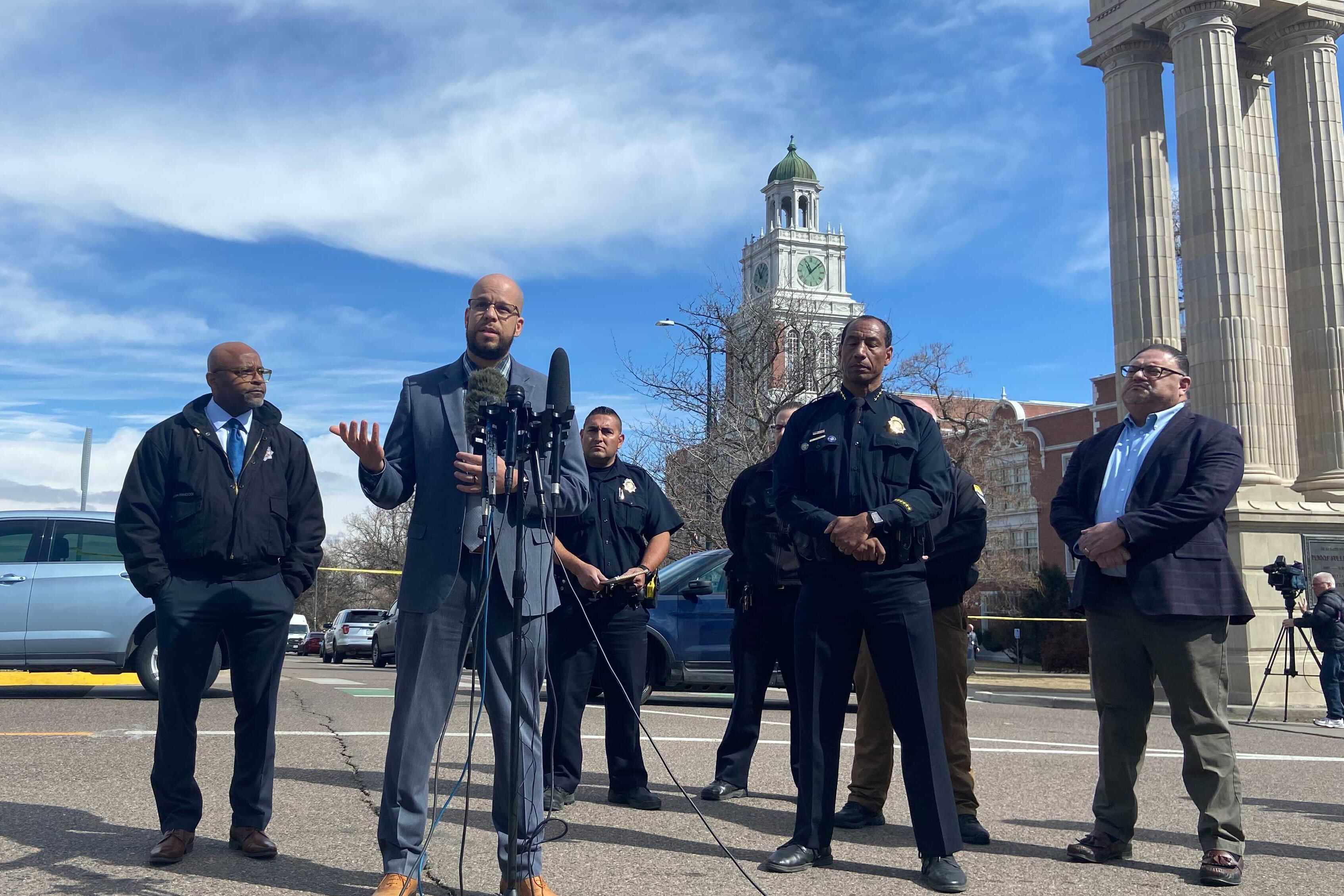 A group of men in suits and one in police dress uniform speak at a press conference. They are outside with a podium and microphones set up in front of them. East High School is visible behind them.