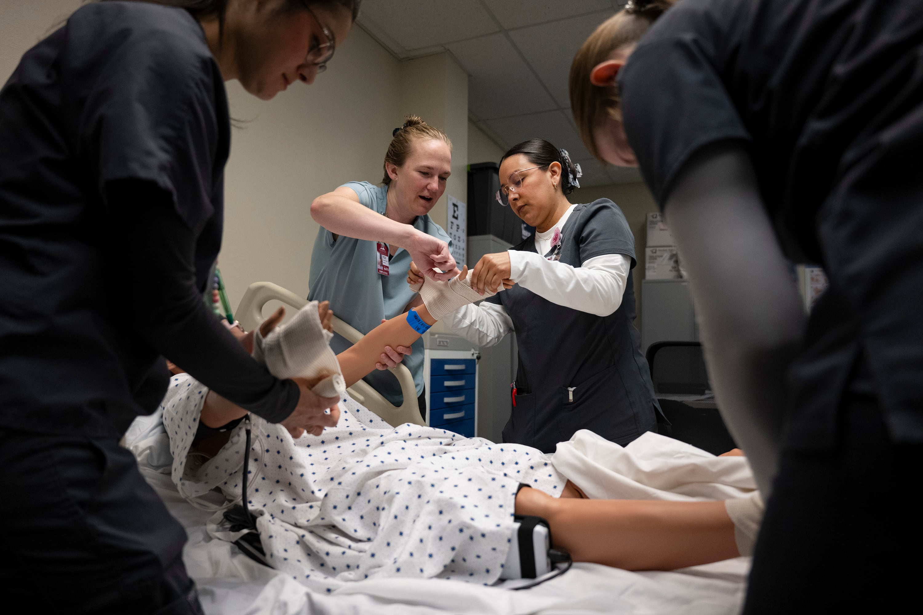 Four females in scrubs are gathered around a treatment bed, wrapping a training manikin's wrist.