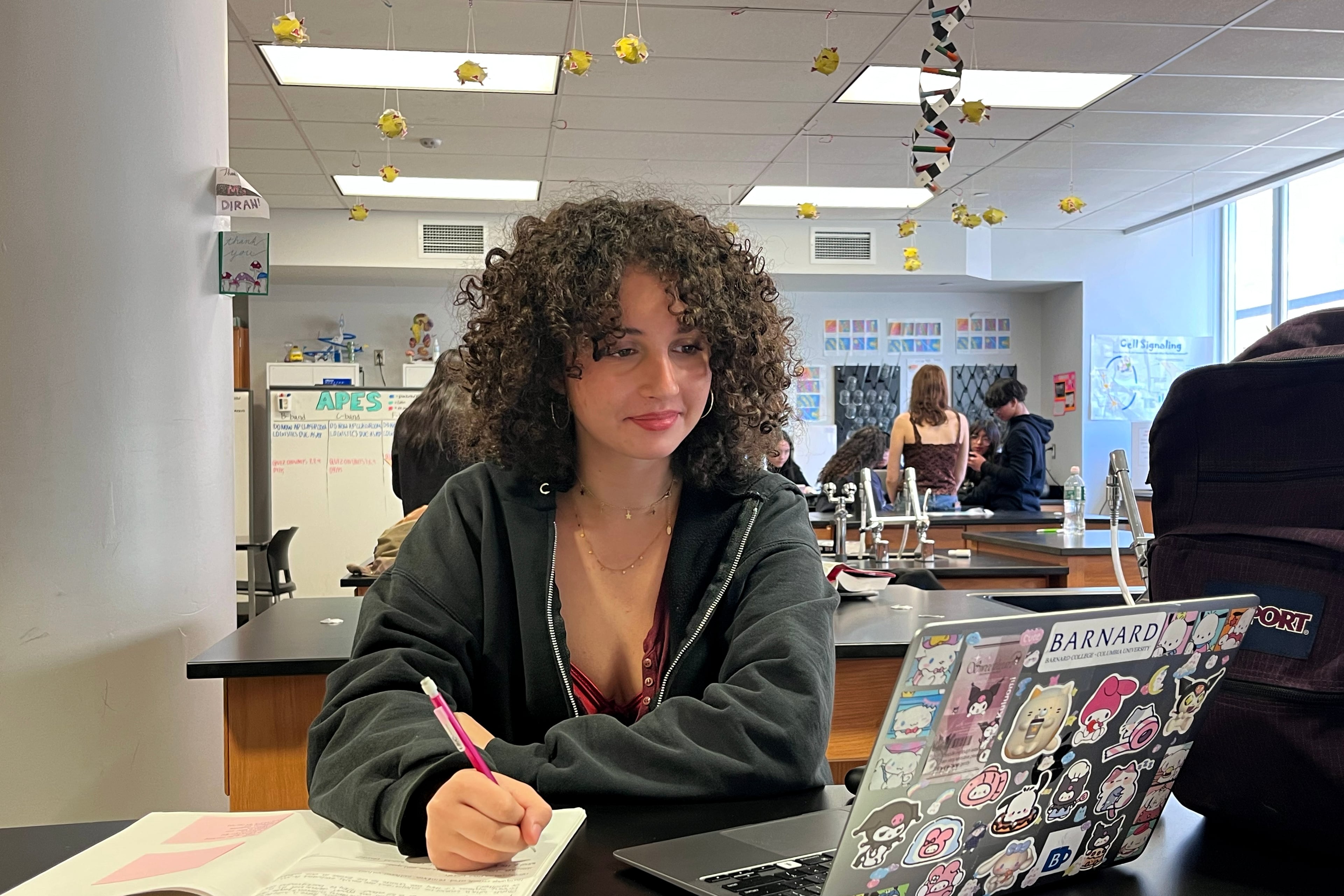 A high school student works at a desk writing in a notebook and looking at a laptop with other students in the background.