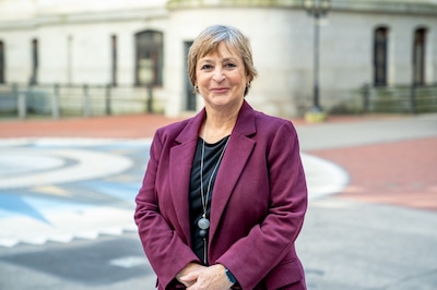 A woman with light hair stands in a maroon jacket and dark top.