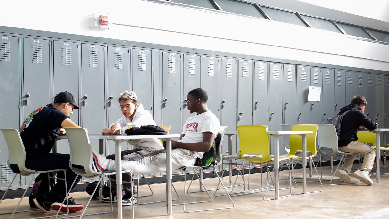 Students sit at small tables together, set against rows of grey lockers.