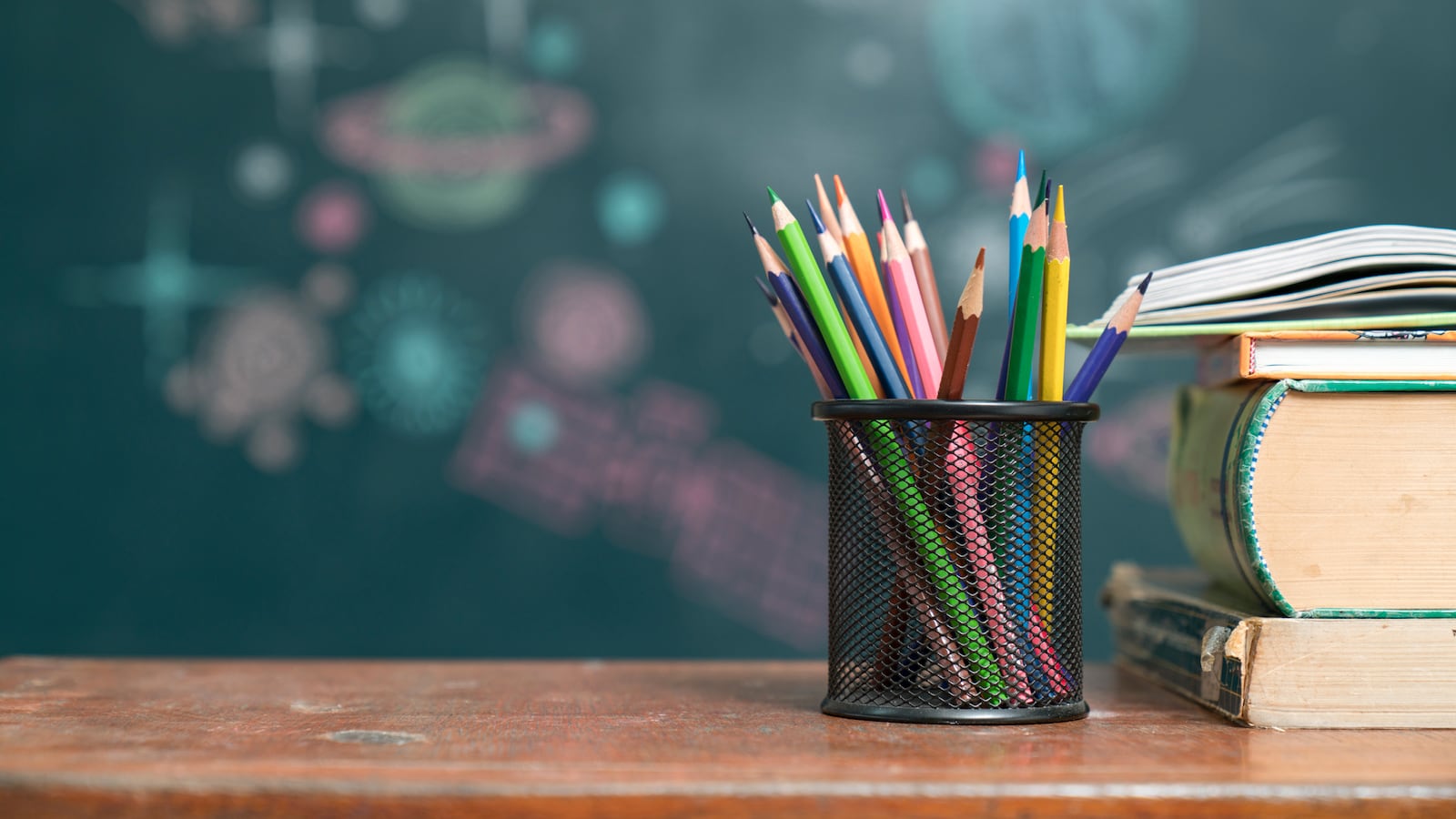 A container with multicolored pencils and a stack of books sit on a desk.
