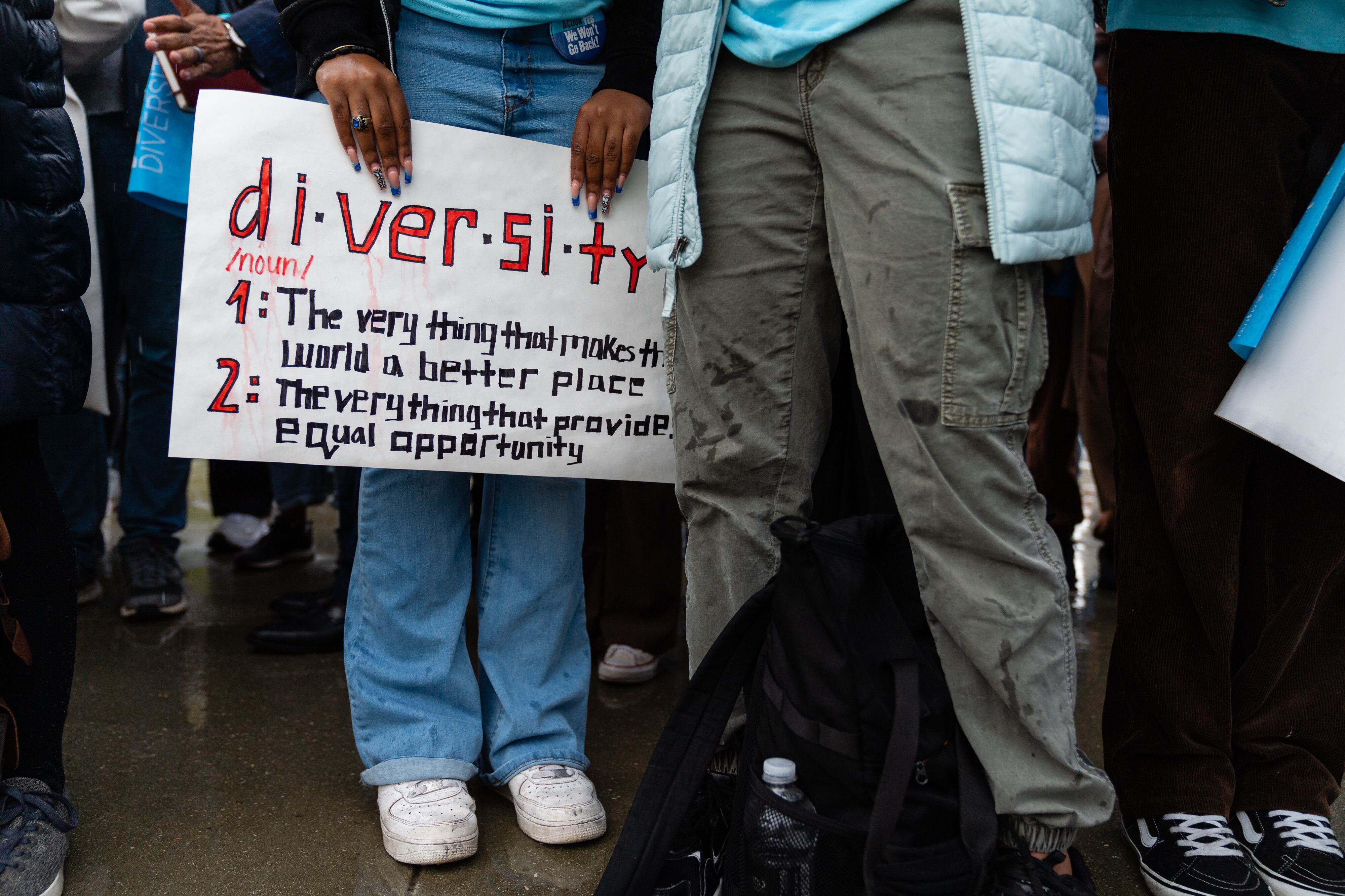 A Black girl wearing jeans and white sneakers holds a sign that says "diversity."