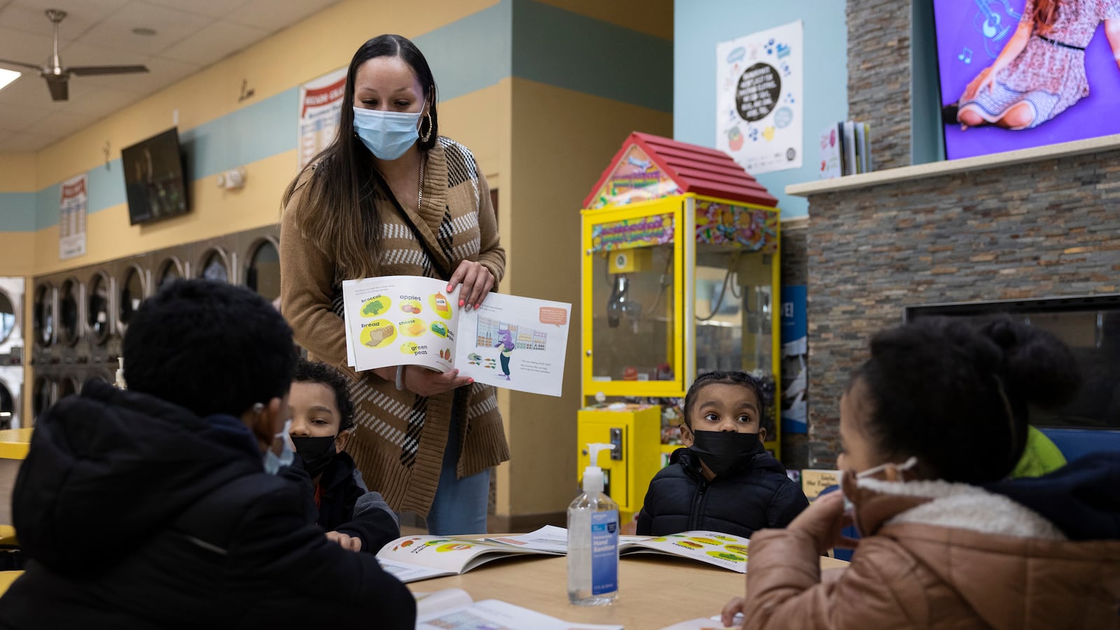 A woman holds a copy of a children’s book toward a group of young readers sitting at a small wooden desk. They are working together in a laundromat.