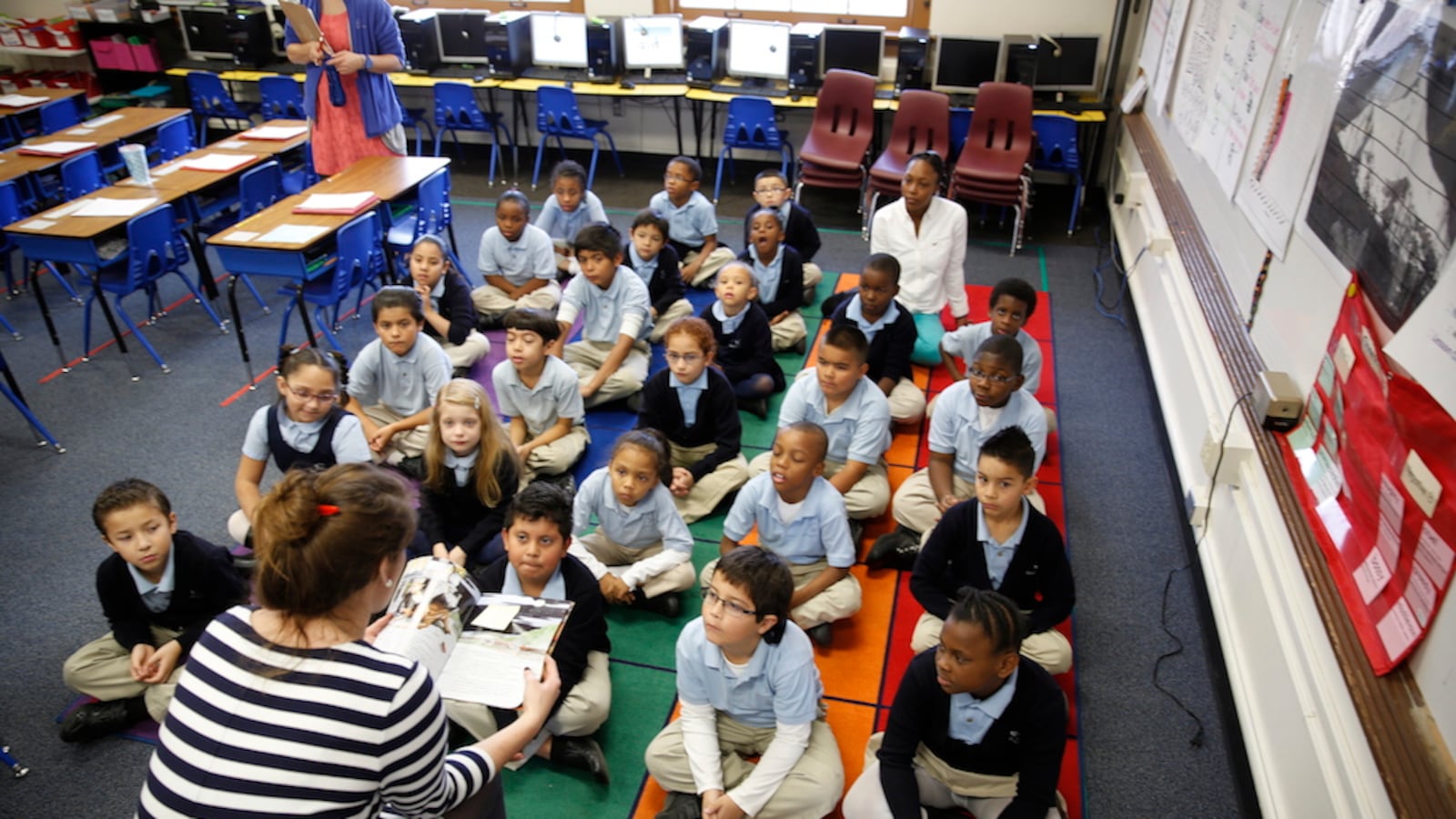 Young students in matching school uniforms sit criss-cross-applesauce on a colorful rug. They are facing the camera. A teacher with her back to the camera reads the students a book.