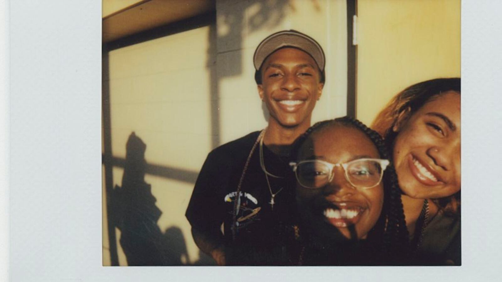 Three teens smile as they face the camera. One of the teen women is wearing glasses. They are framed by a shadow of a window.