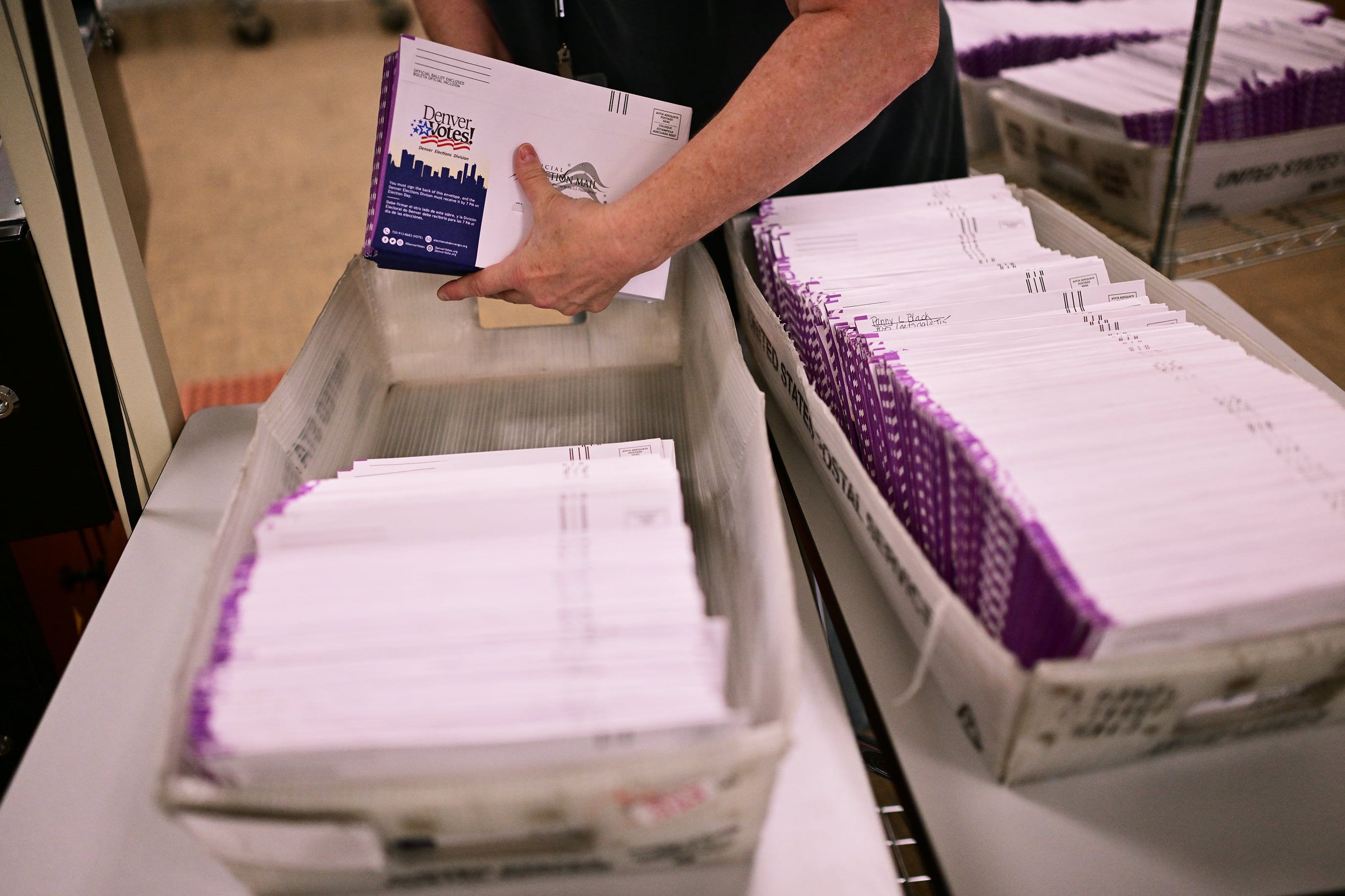 A photograph of a stack of white mail in ballot envelopes on a wire rack and an up close view of a person's arm carrying a stack of envelopes.