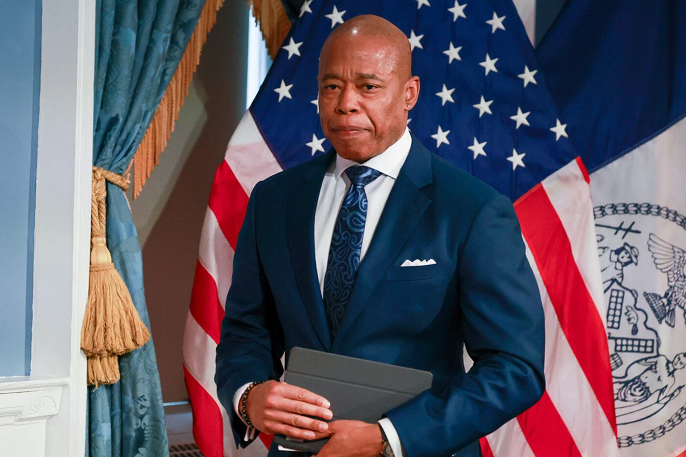 A man wearing a dark blue suit holds a black binder and stands in front of an American flag.