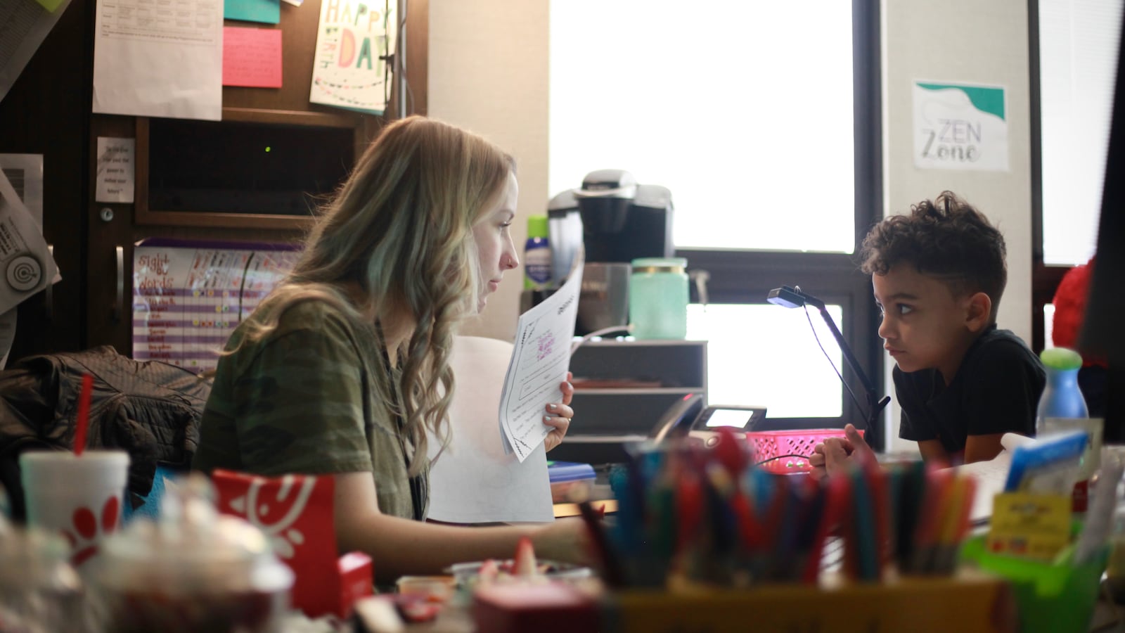A young teacher holds up a paper as she speaks with a boy at her desk.