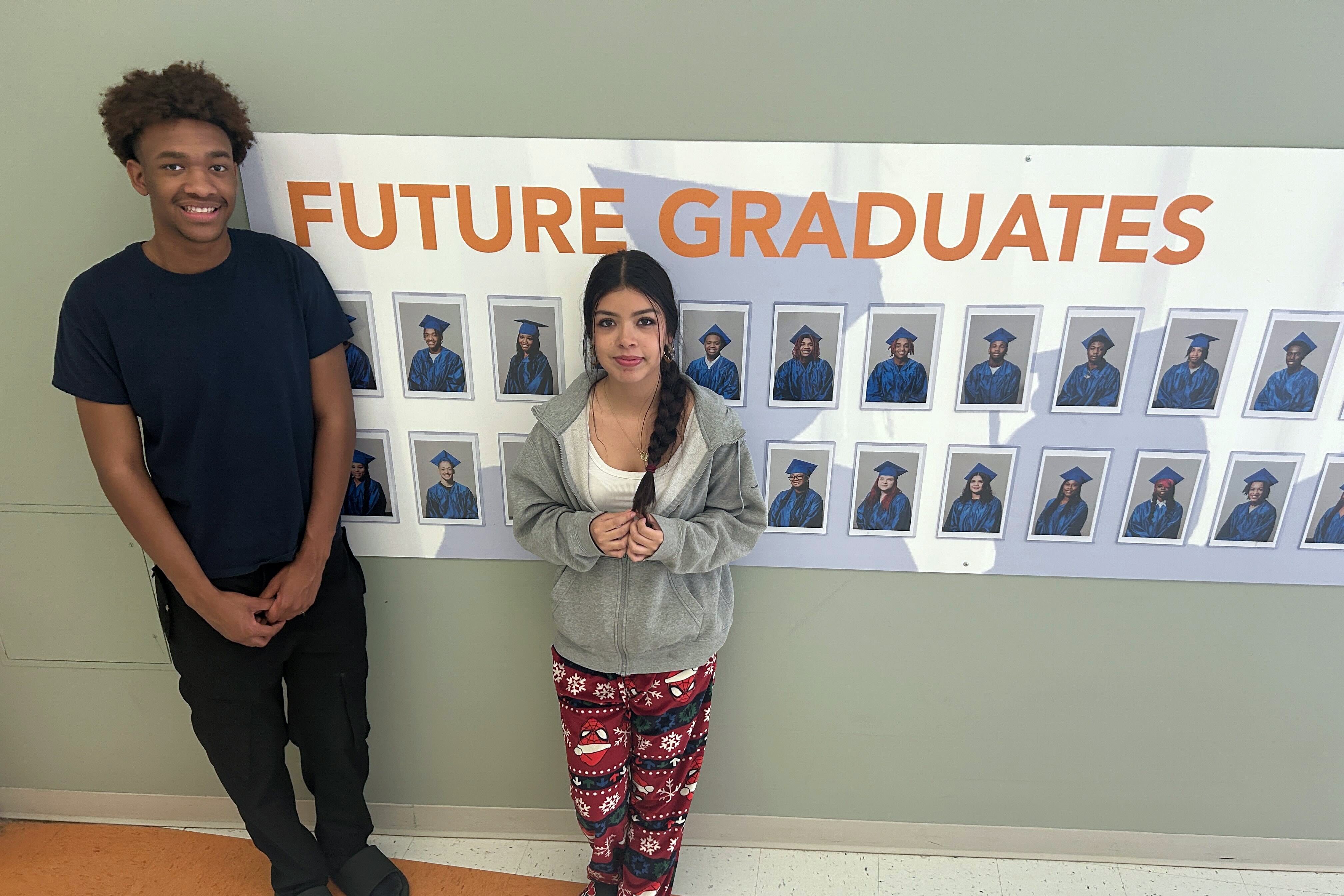 Two young high school students stand next to a wall with a poster of portraits of students and a title that reads "Future Graduates."