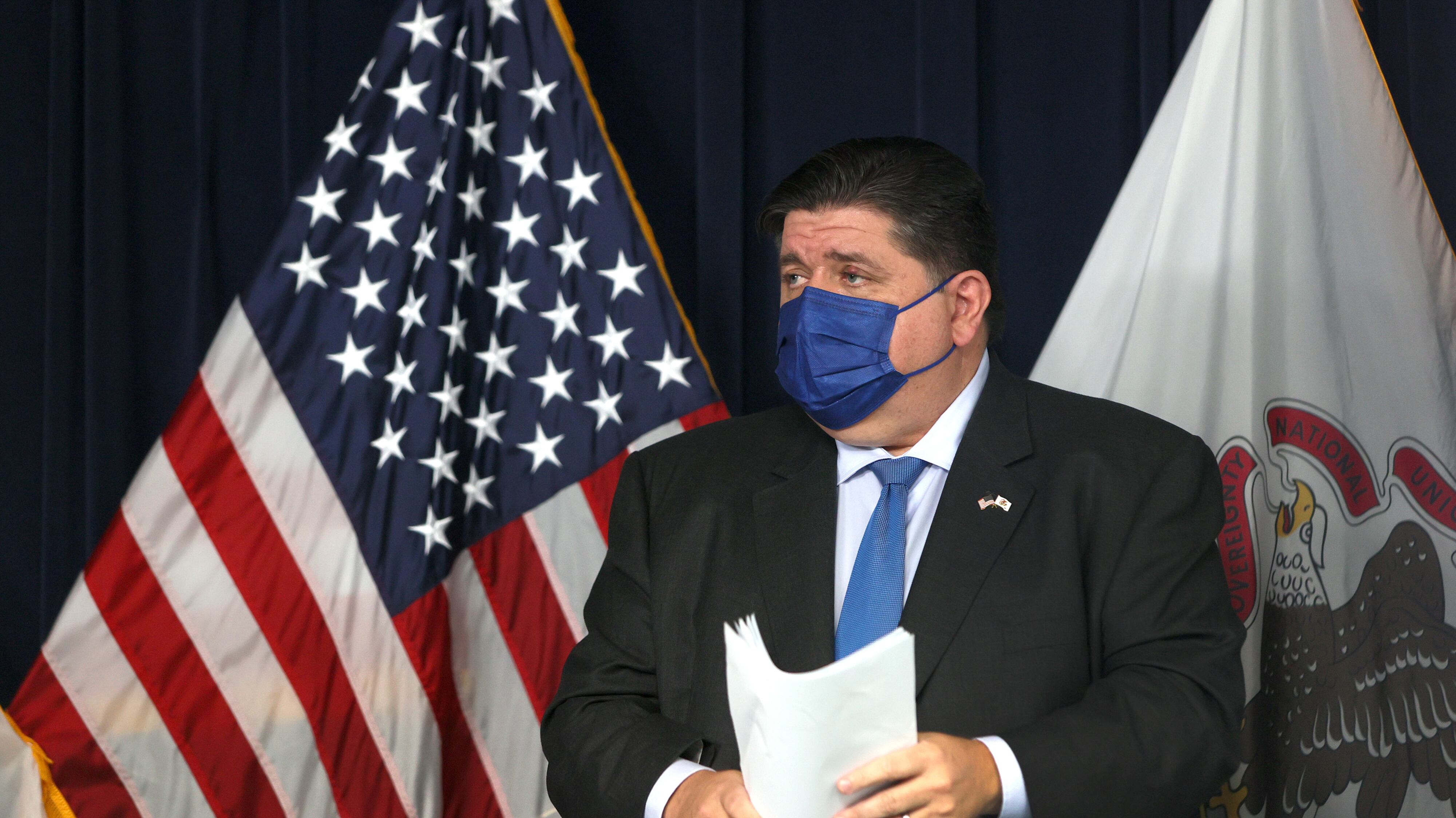 Governor J.B. Pritzker holds papers and stands on a stage in front of the United States flag, wearing a blue mask.