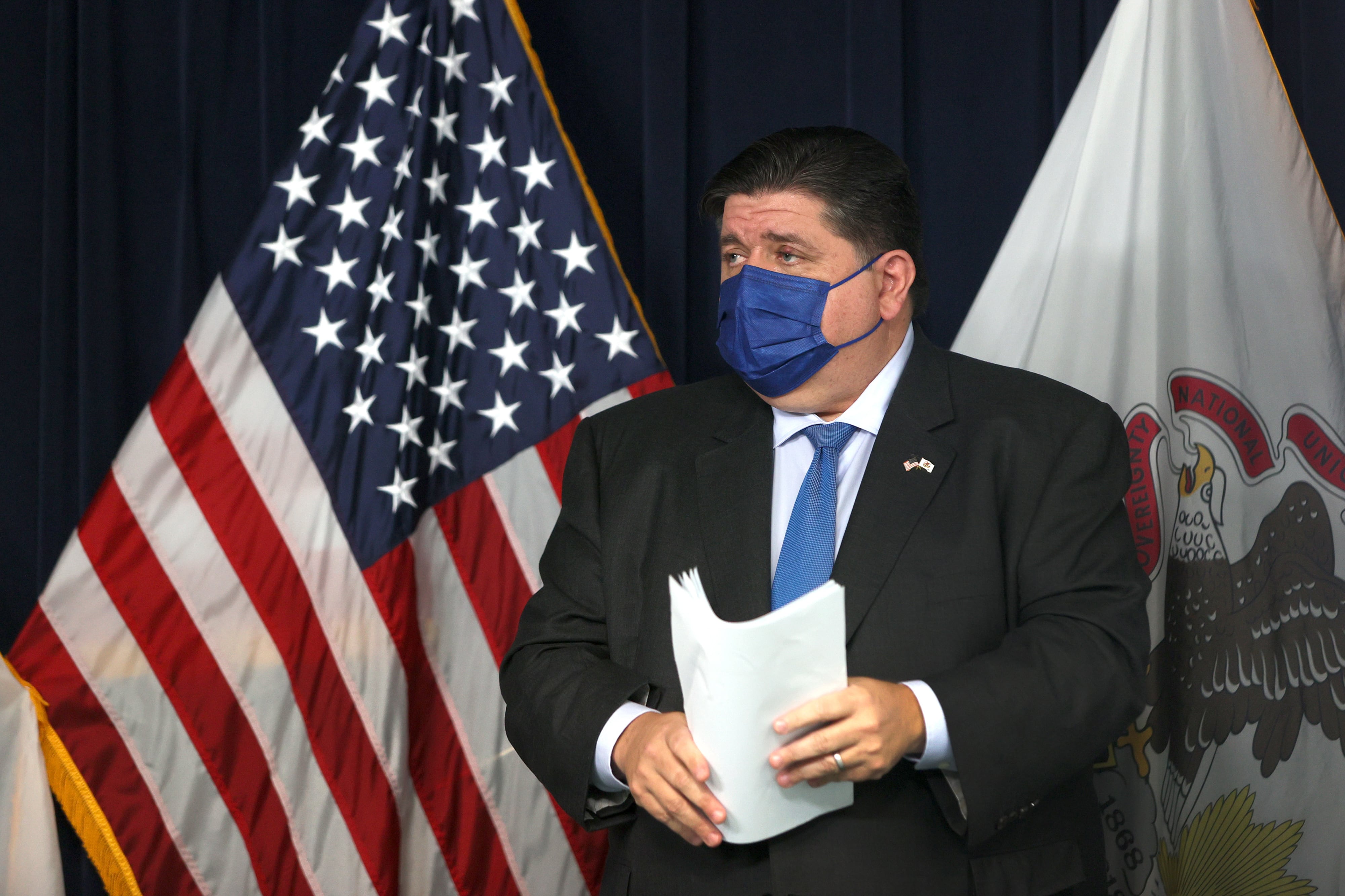 Governor J.B. Pritzker holds papers and stands on a stage in front of the United States flag, wearing a blue mask.