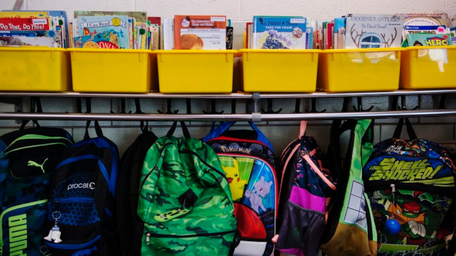 Books in yellow bins sit on a shelf and backpacks hang from hooks in a classroom.
