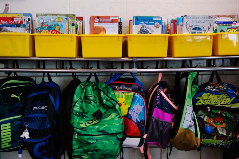 Books in yellow bins sit on a shelf and backpacks hang from hooks in a classroom.