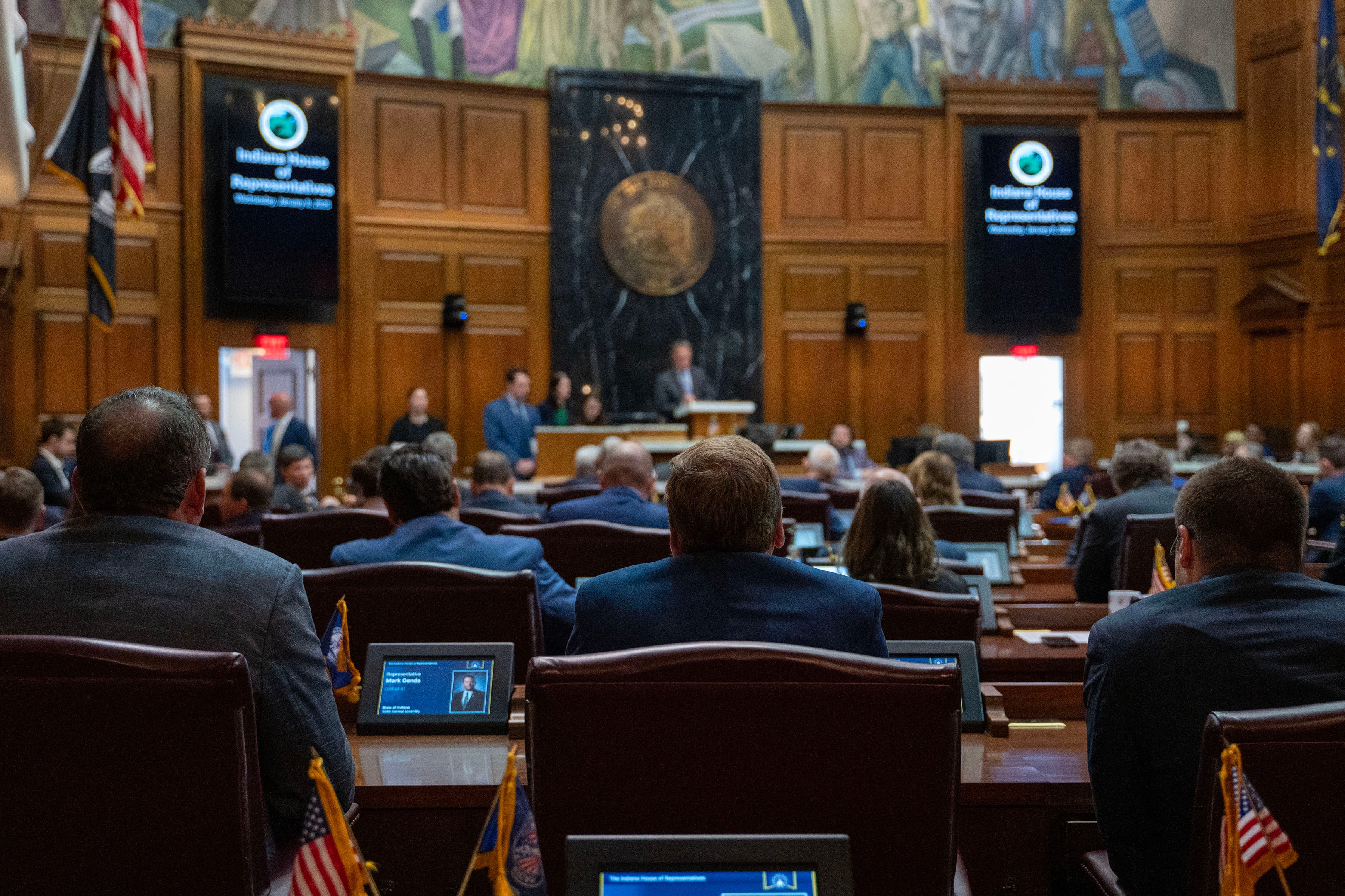 A room full of people in business clothes sit at chairs in rows of wooden desks in a large room.