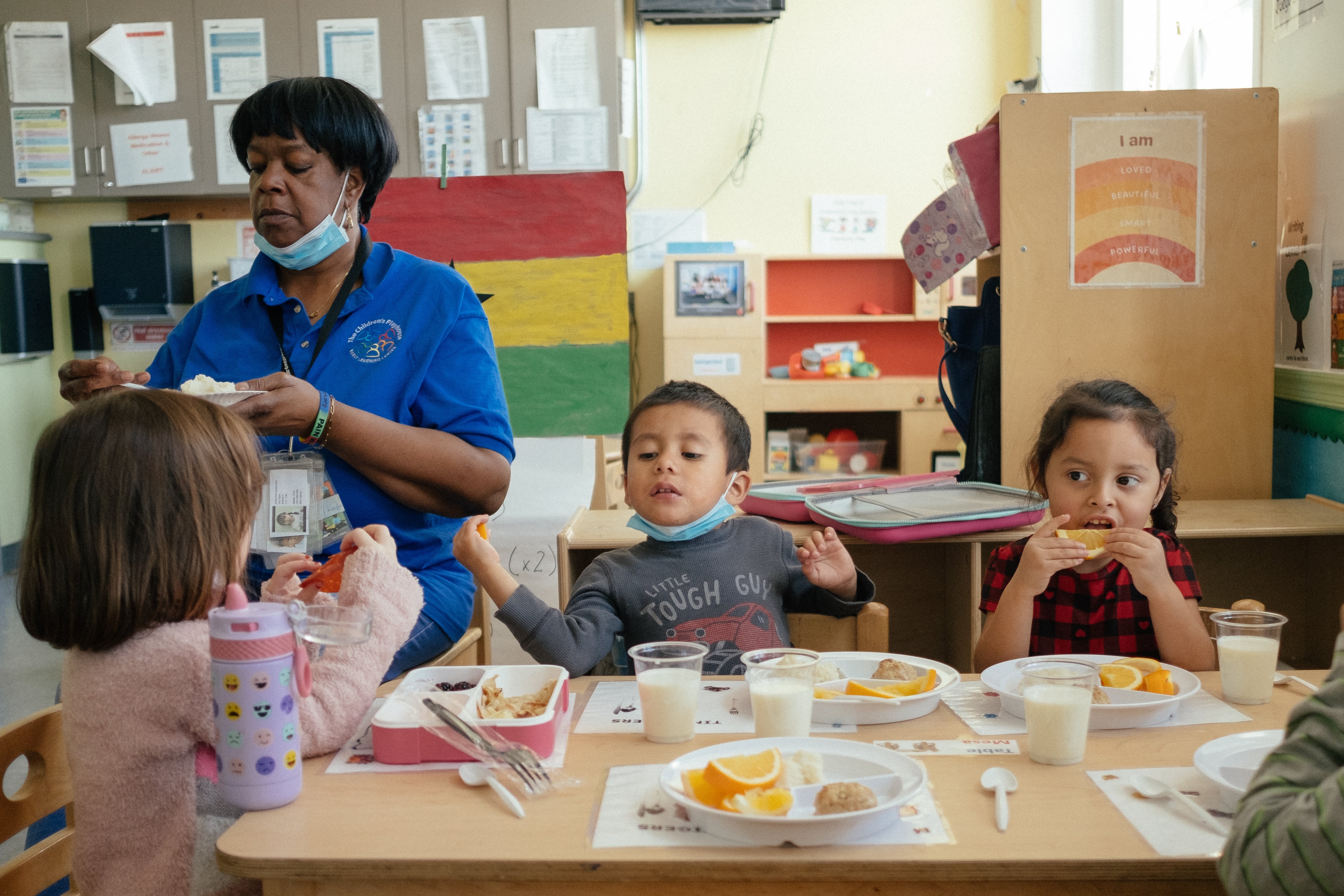Students eat lunch with one of their teachers at a small wooden table.