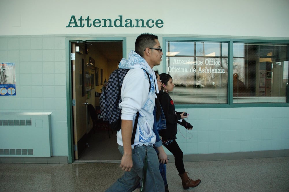Three high school students walk past a light and dark green brick wall with the words "Attendance" near the top.