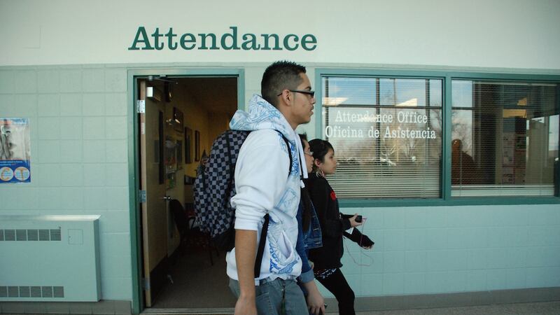 Three high school students walk past a light and dark green brick wall with the words "Attendance" near the top.