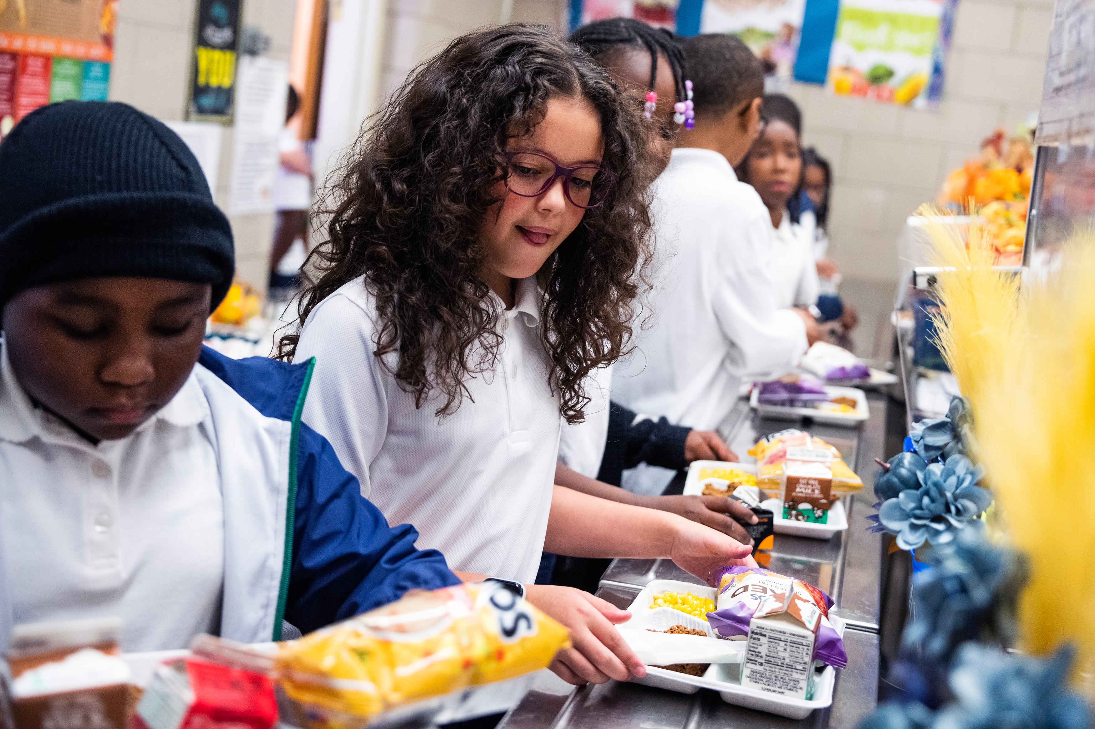 A row of elementary students all wearing white shirts, stand in line at a cafeteria while holding a tray of food.