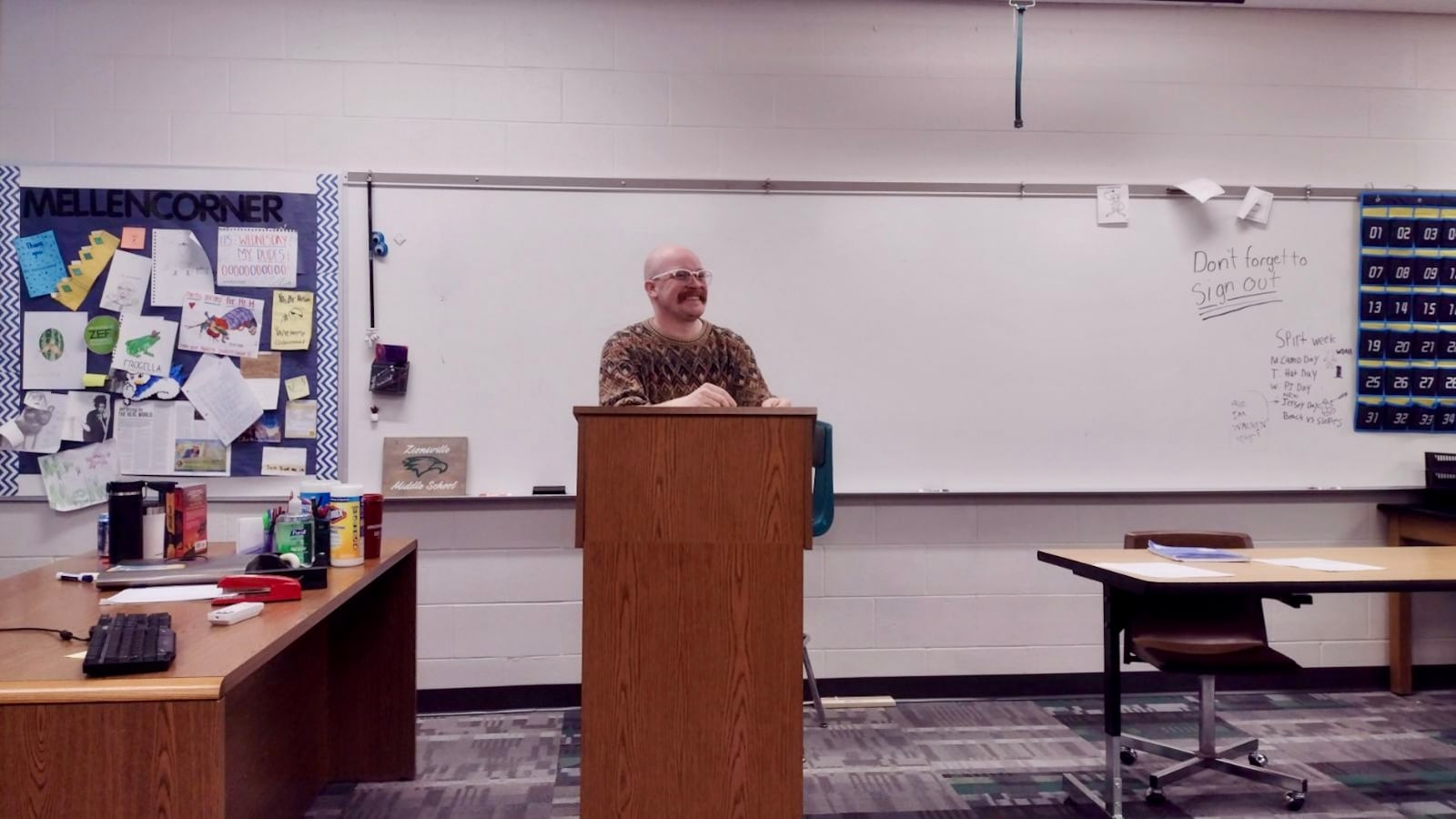 A man stands behind a wooden podium in a classroom. A white board is behind him.