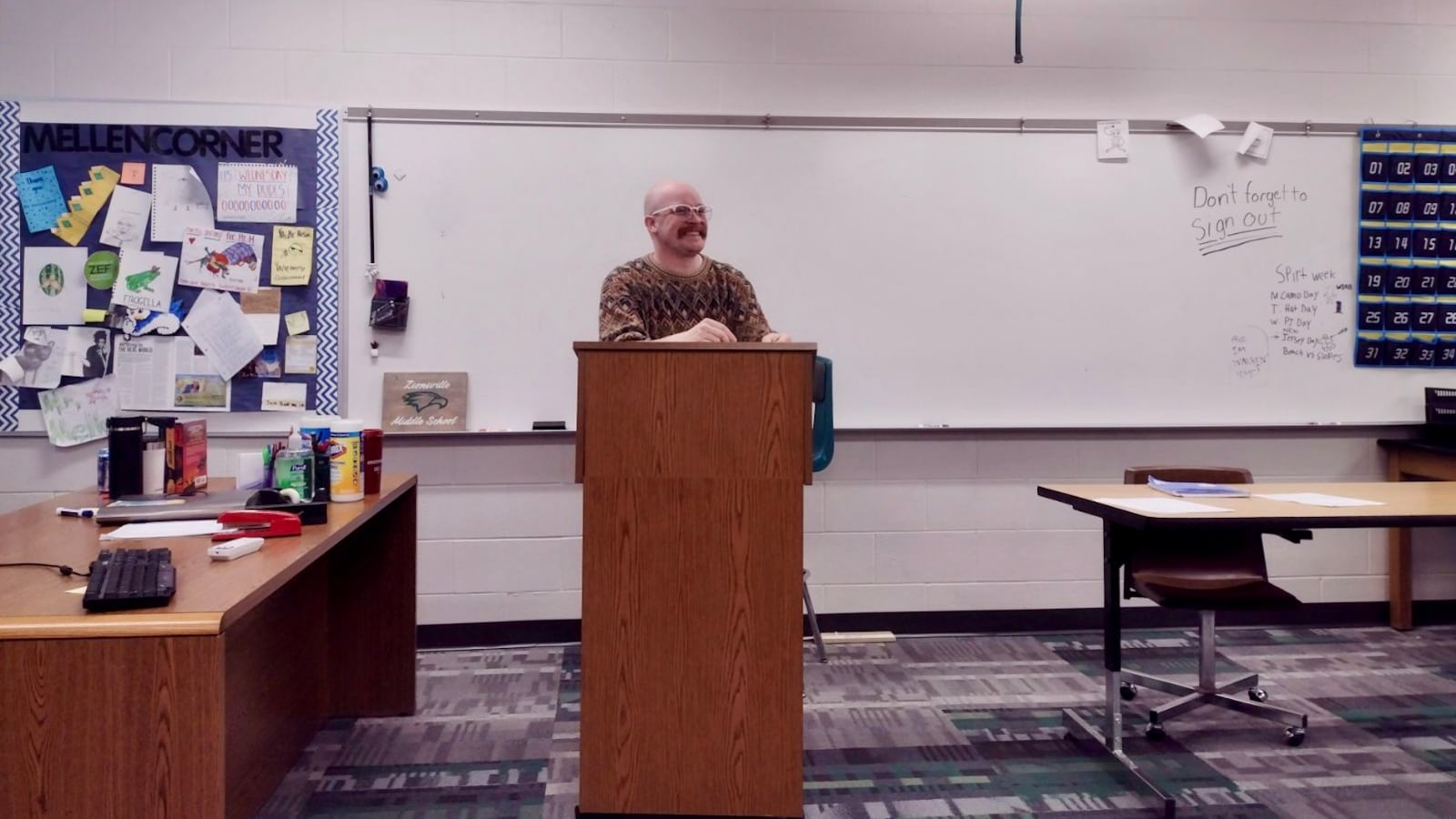 A man stands behind a wooden podium in a classroom. A white board is behind him.