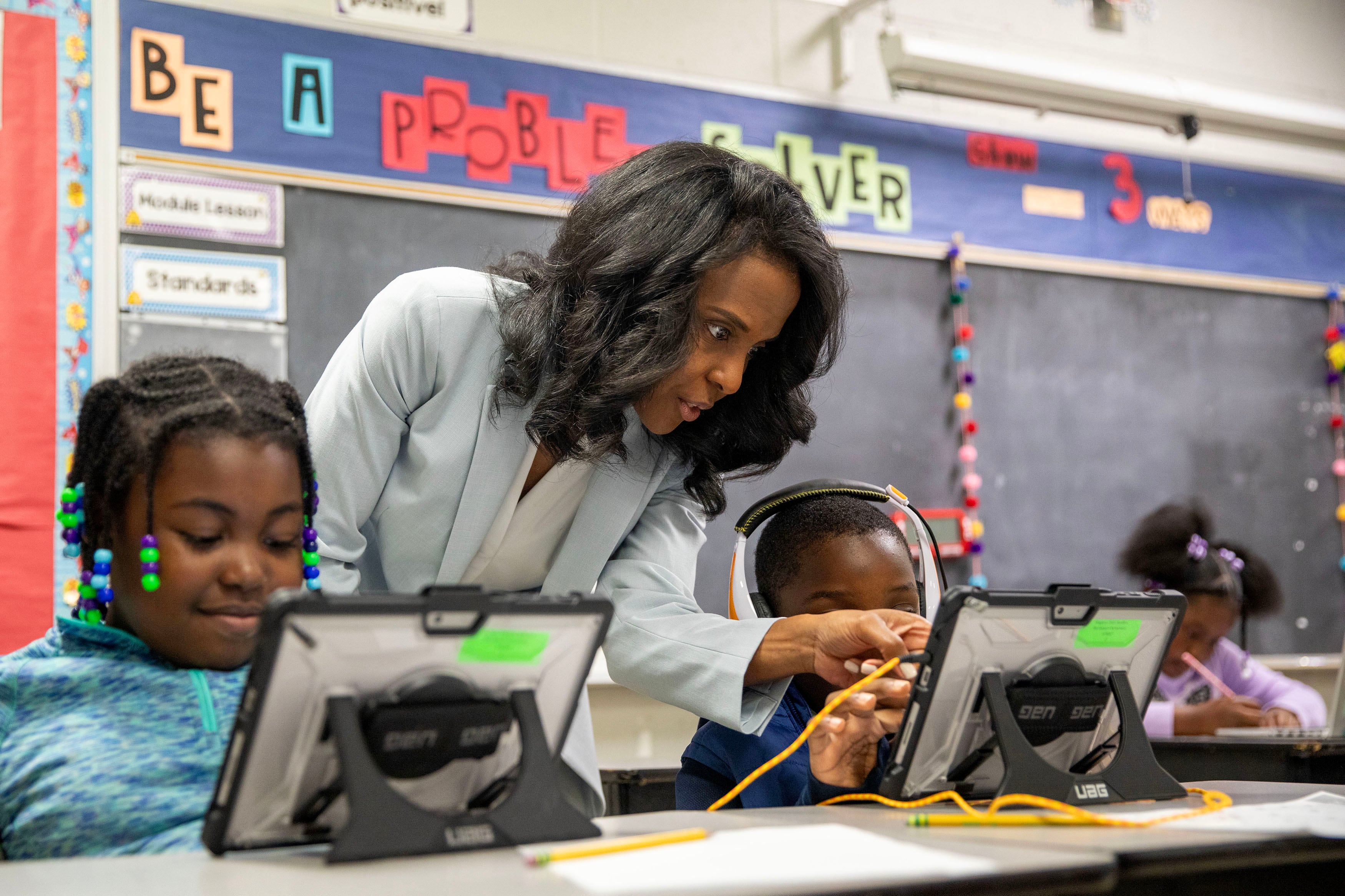 An adult in a blue suit stands next to two young students working at a desk in a classroom.