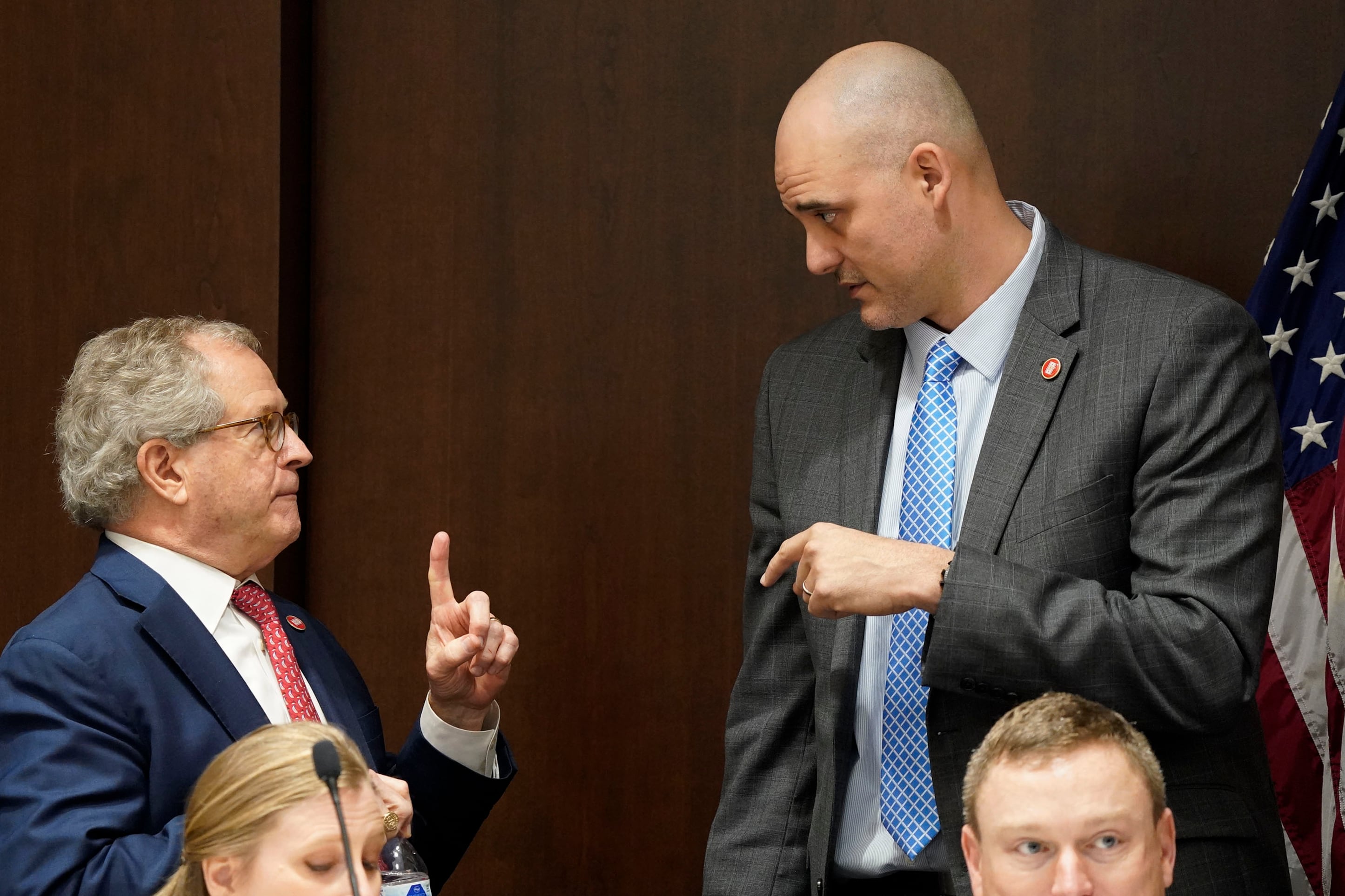 Two men wearing business suits speak with each other in a committee room.