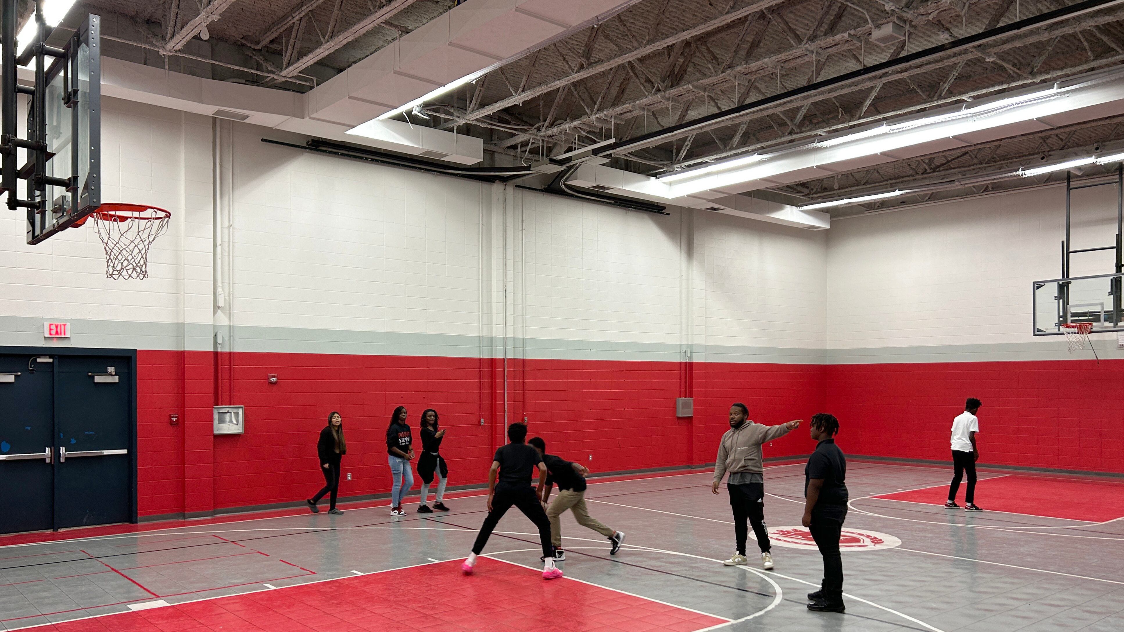 Eight people play basketball or stand in a gym with red and grey flooring and tall walls.