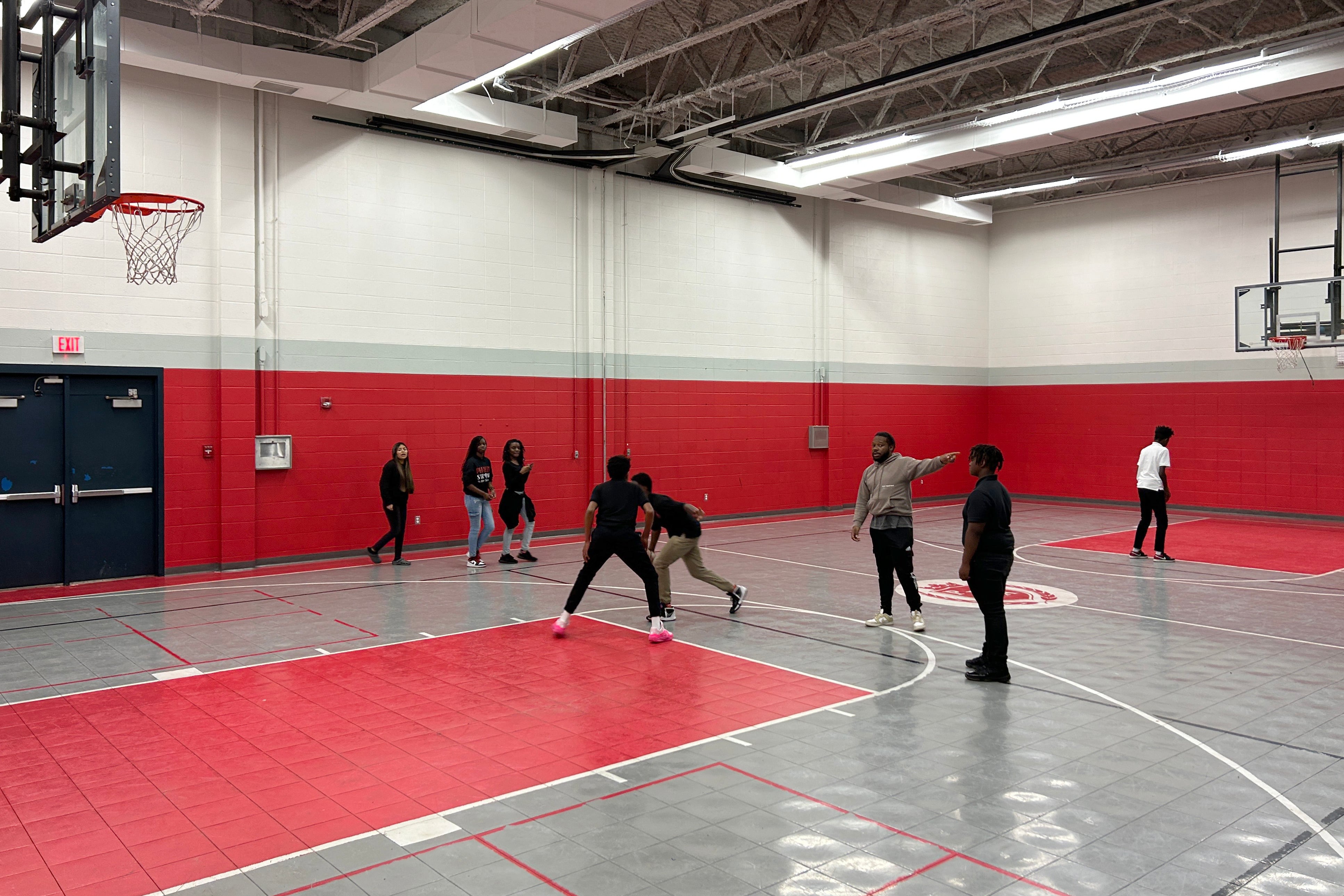 Eight people play basketball or stand in a gym with red and grey flooring and tall walls.