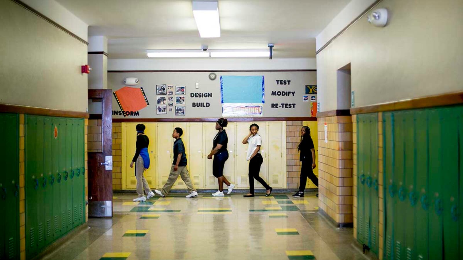 Students walk down a hallway during class change. The walls are lined with green and yellow lockers.