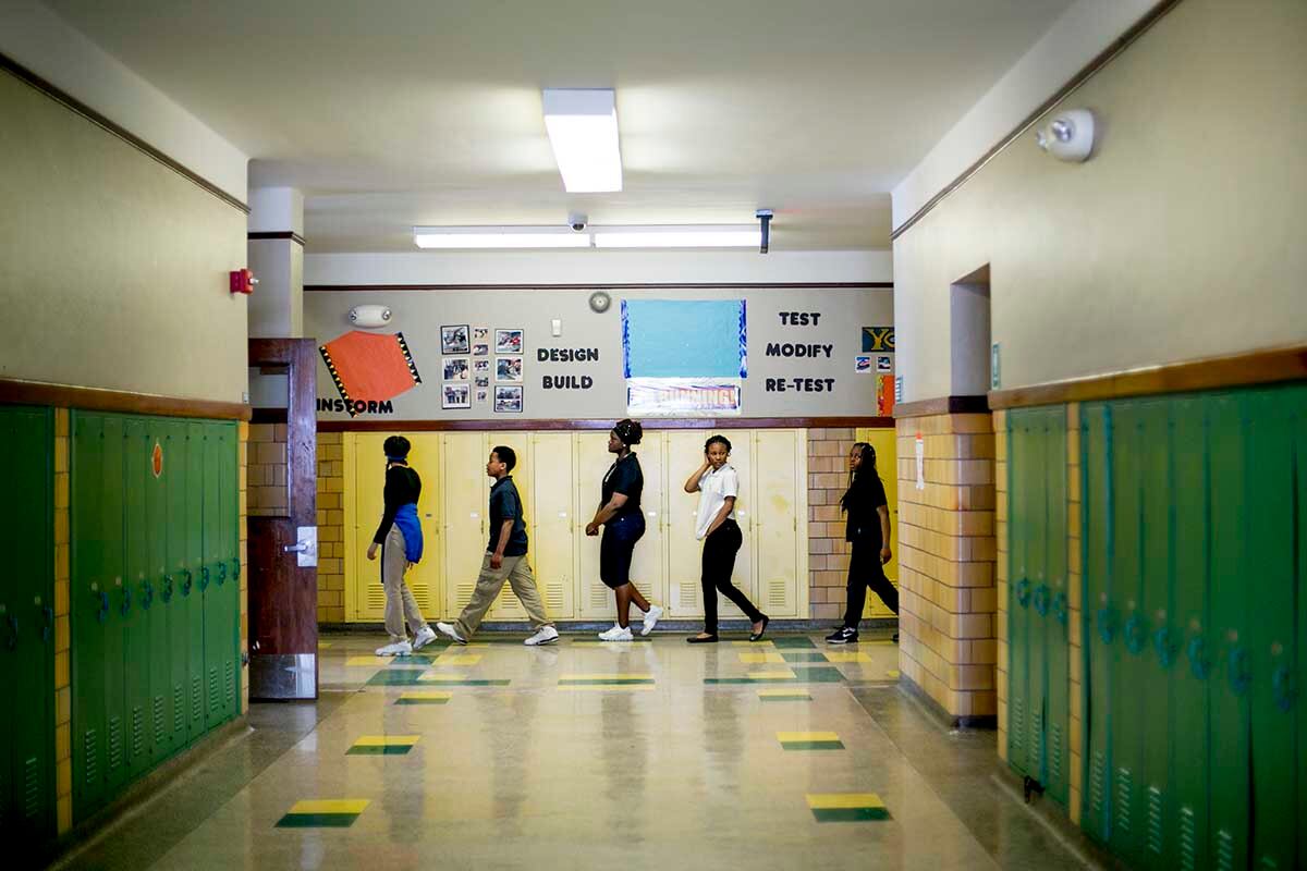 Students walk down a hallway during class change. The walls are lined with green and yellow lockers.