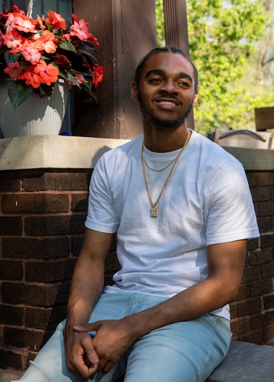 A high school senior with dark hair and wearing a white t-shirt with a gold necklace with a diamond letter "K" poses for a portrait in front of brick wall.