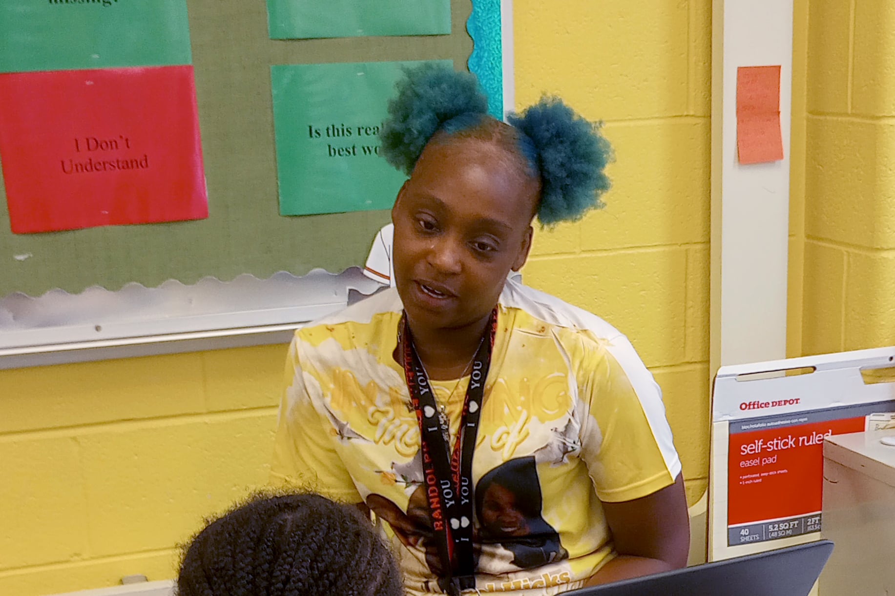 A woman in a yellow T-shirt sits in front of a laptop with a student.