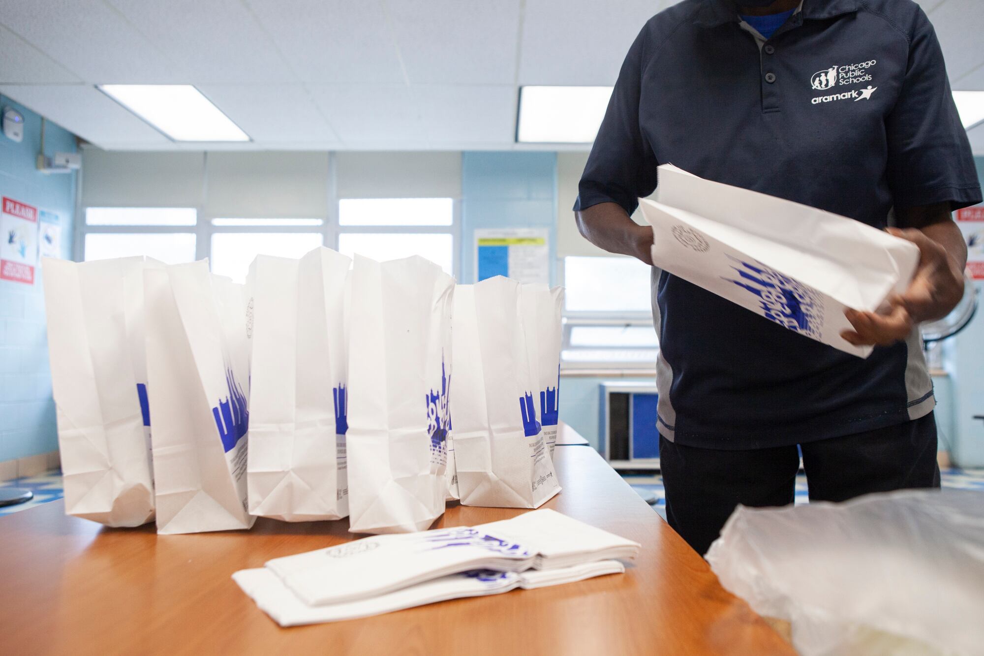 A food service worker in dark color clothes assembles lunch bags in a white classroom on a brown table.