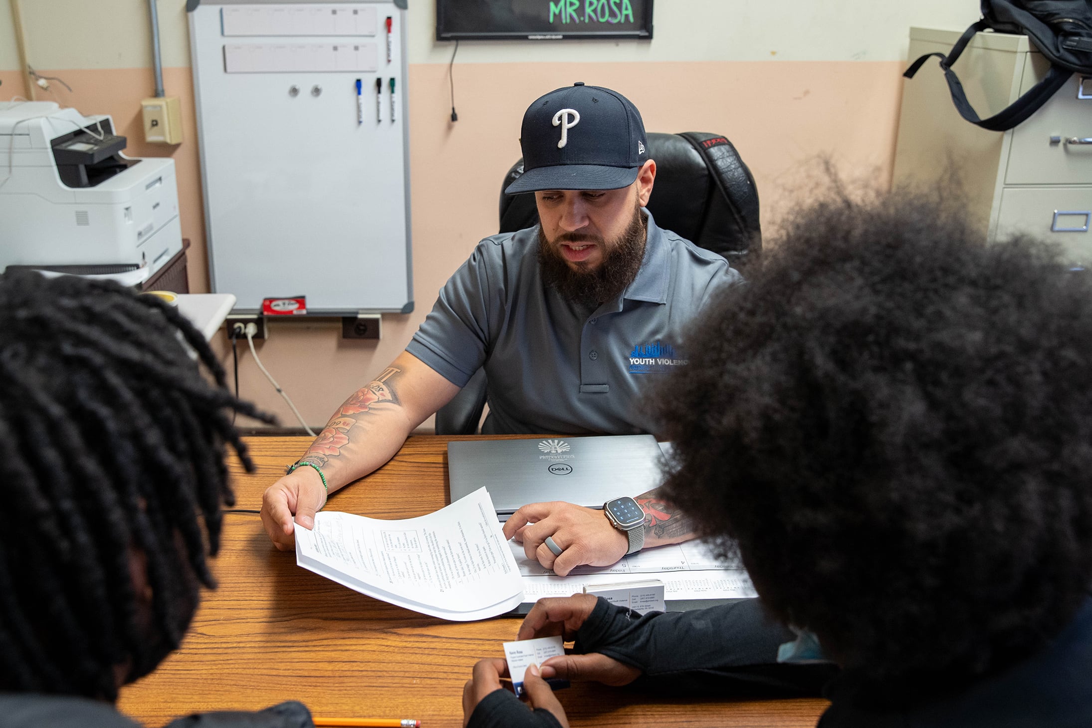 A man wearing a baseball cap and a blue shirt sits behind a wooden desk and talks to two students in a school office.