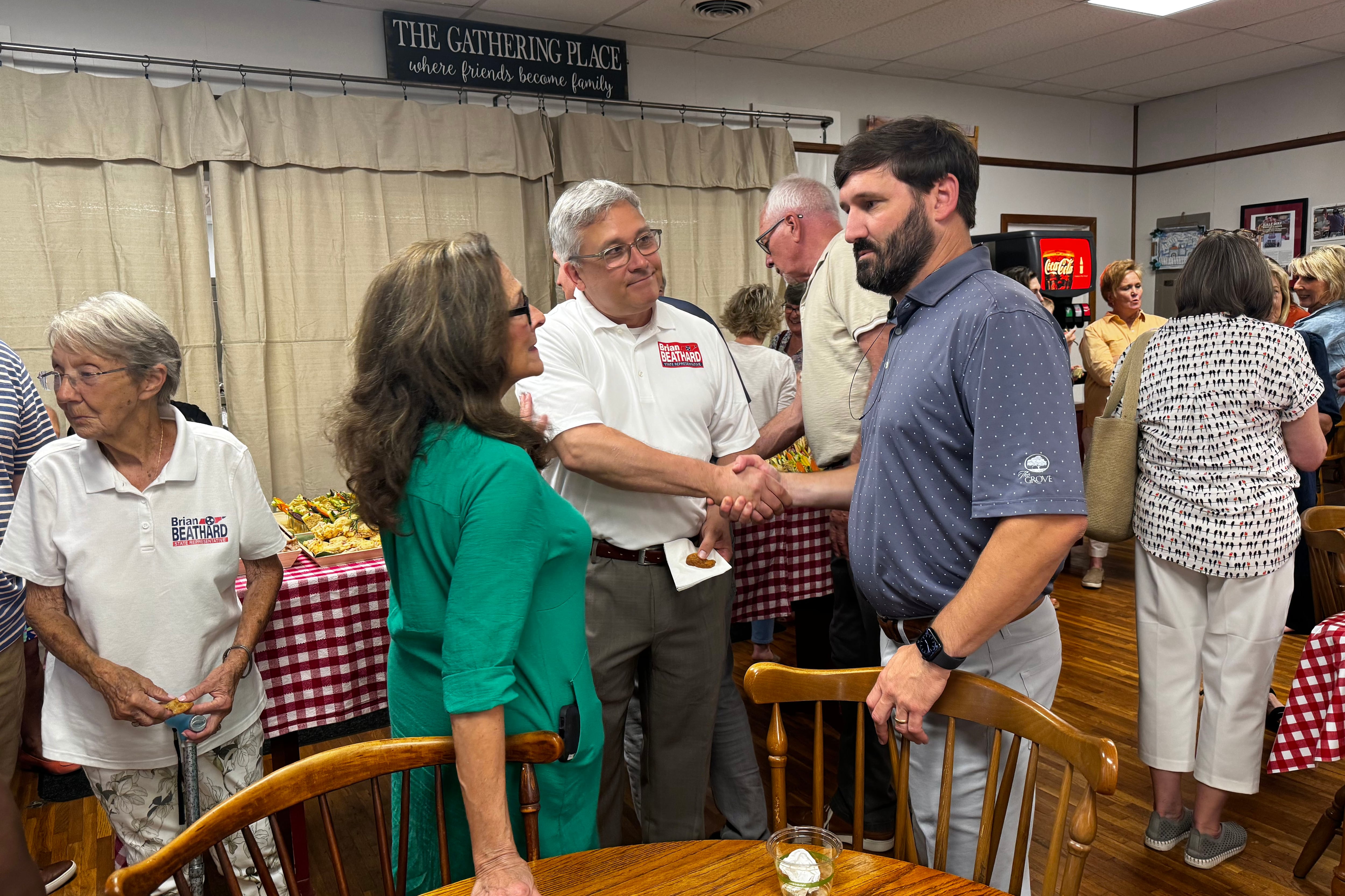 Three people stand next to a wooden table and chairs talking with each other. The person in the center and the left shake hands.