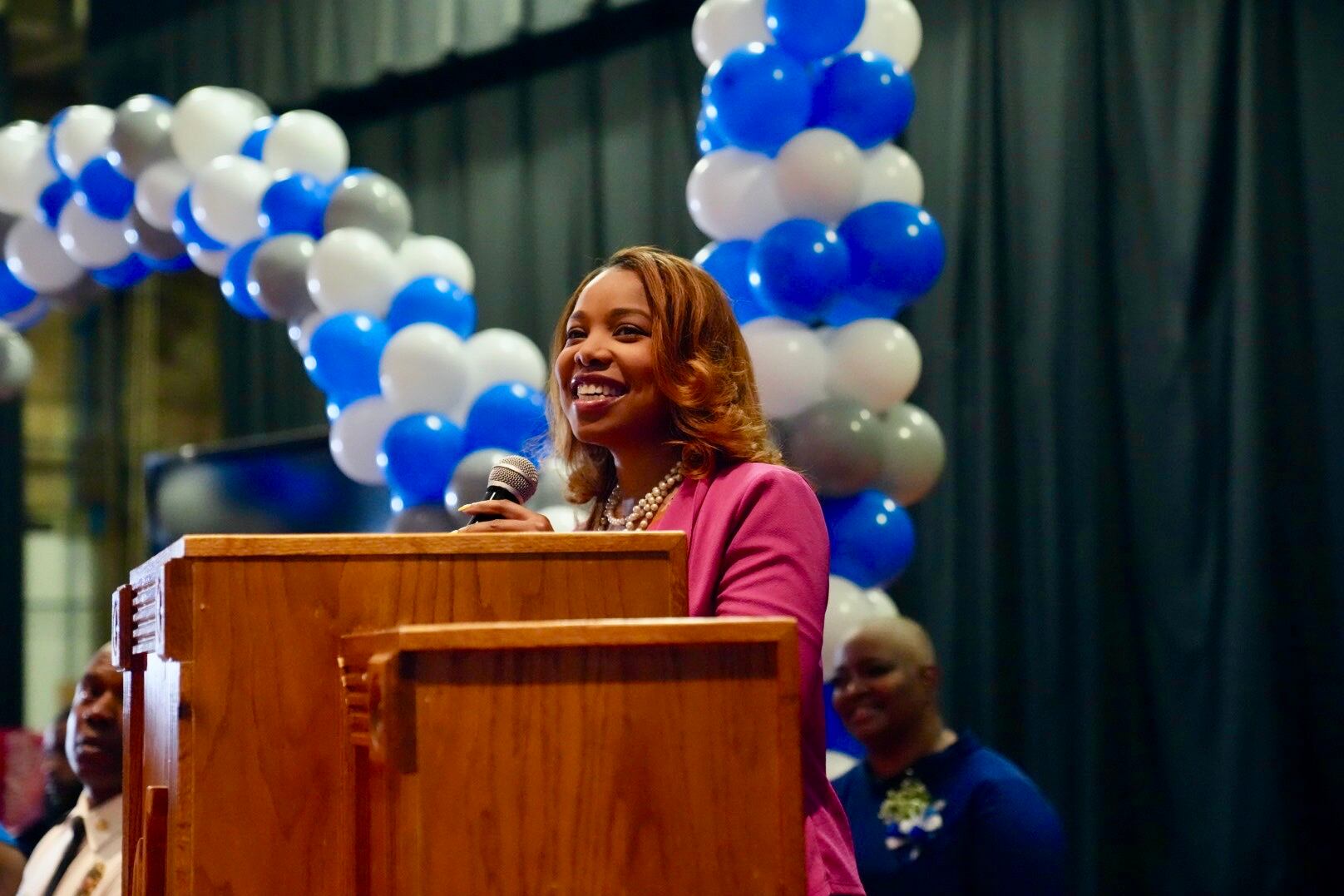A woman standing in front of an arch of blue and white balloons speaks at a podium